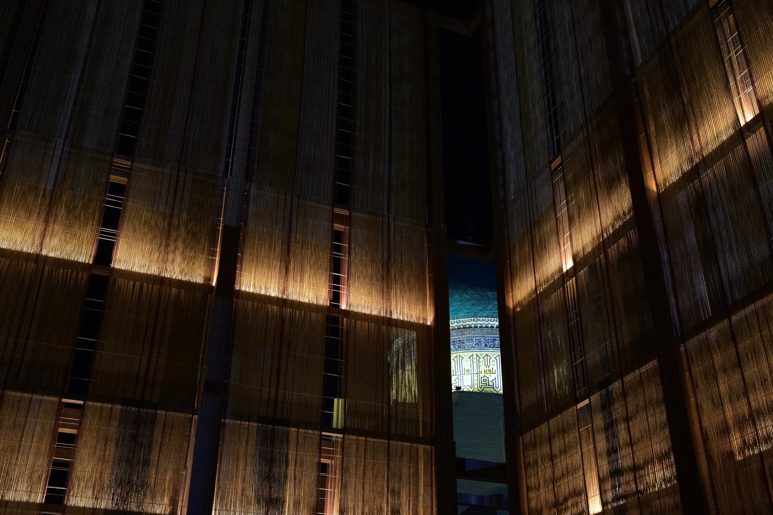 Interior view of a modern building with bamboo or wooden slats on the walls and a brightly lit decorative mosaic-tiled spiral staircase or feature visible through a narrow opening.