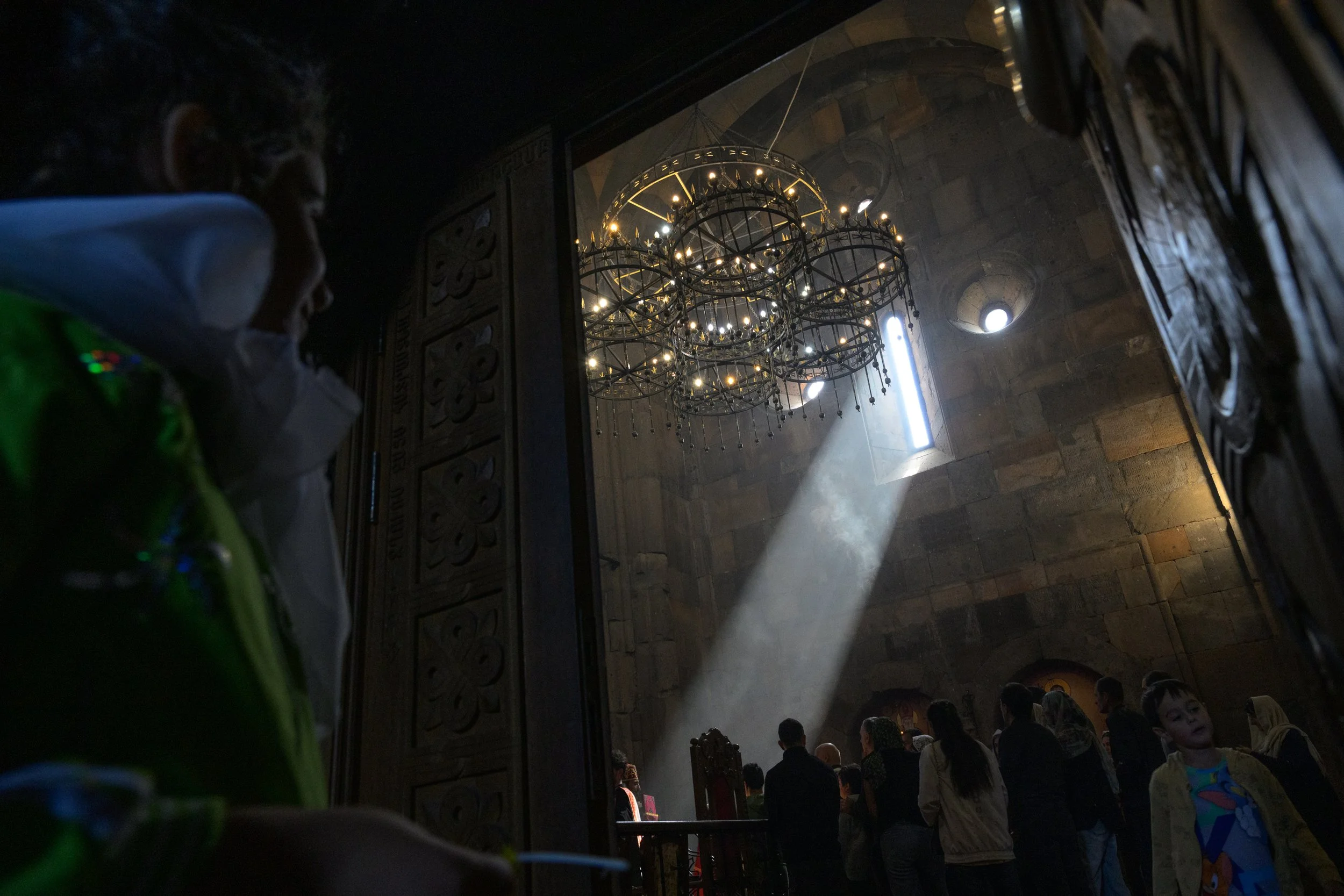 Inside a church or cathedral, with a high ceiling and a large chandelier hanging. Sunlight streams through a vertical window, illuminating a beam of light inside. People are gathered, some standing in line or waiting, with one young boy visible in th