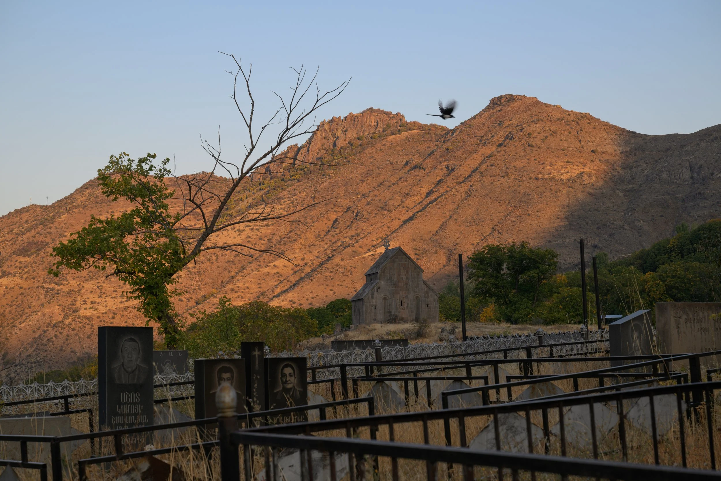 A cemetery with headstones and a small church, set against a mountain landscape at sunset, with a bird flying in the sky, and a leafless tree in the foreground.
