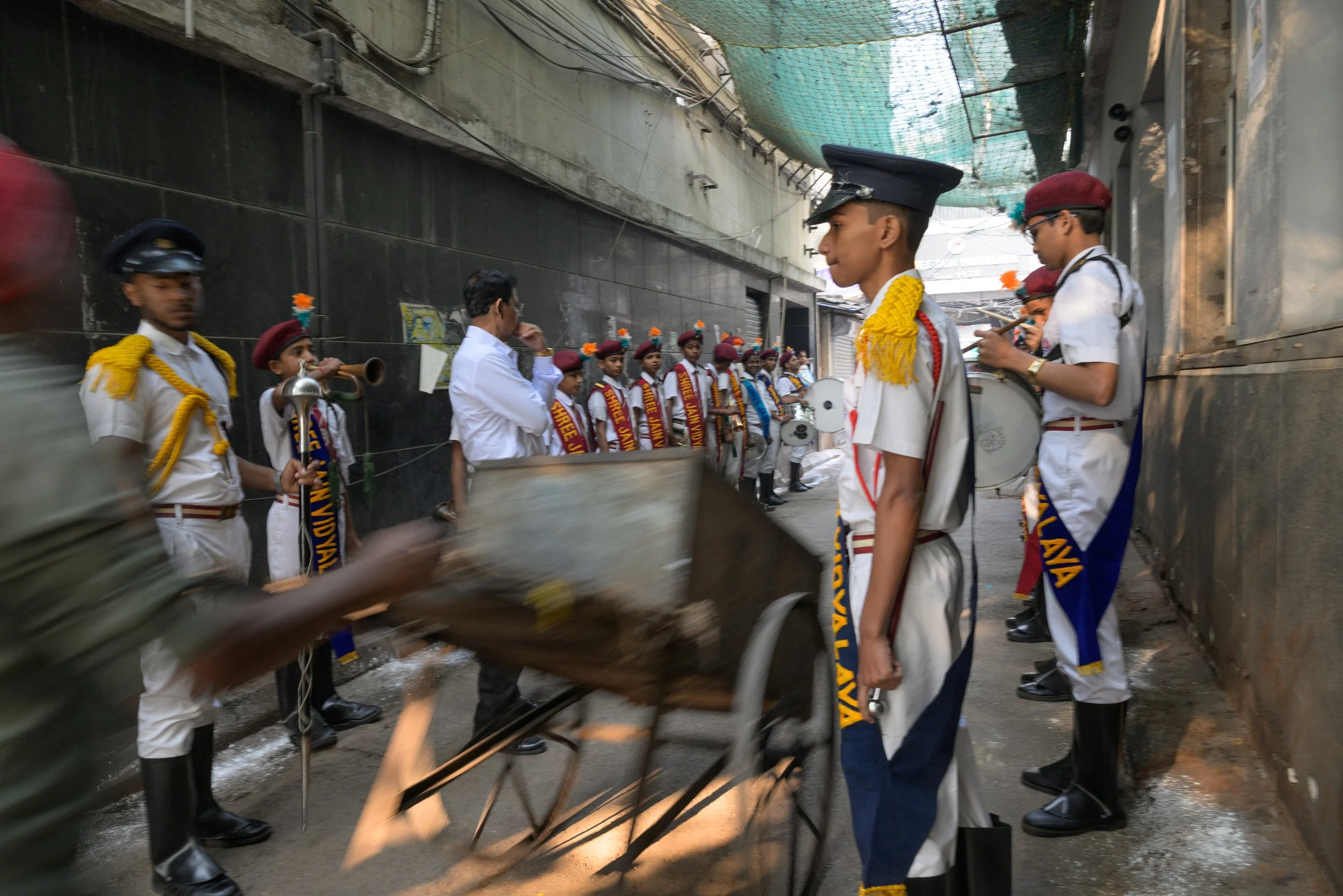 A group of school children dressed in white uniforms with red scarves and maroon hats, some holding musical instruments like drums and a trumpet, are gathered in an alleyway for a street parade or celebration, with a man in a white shirt standing in 