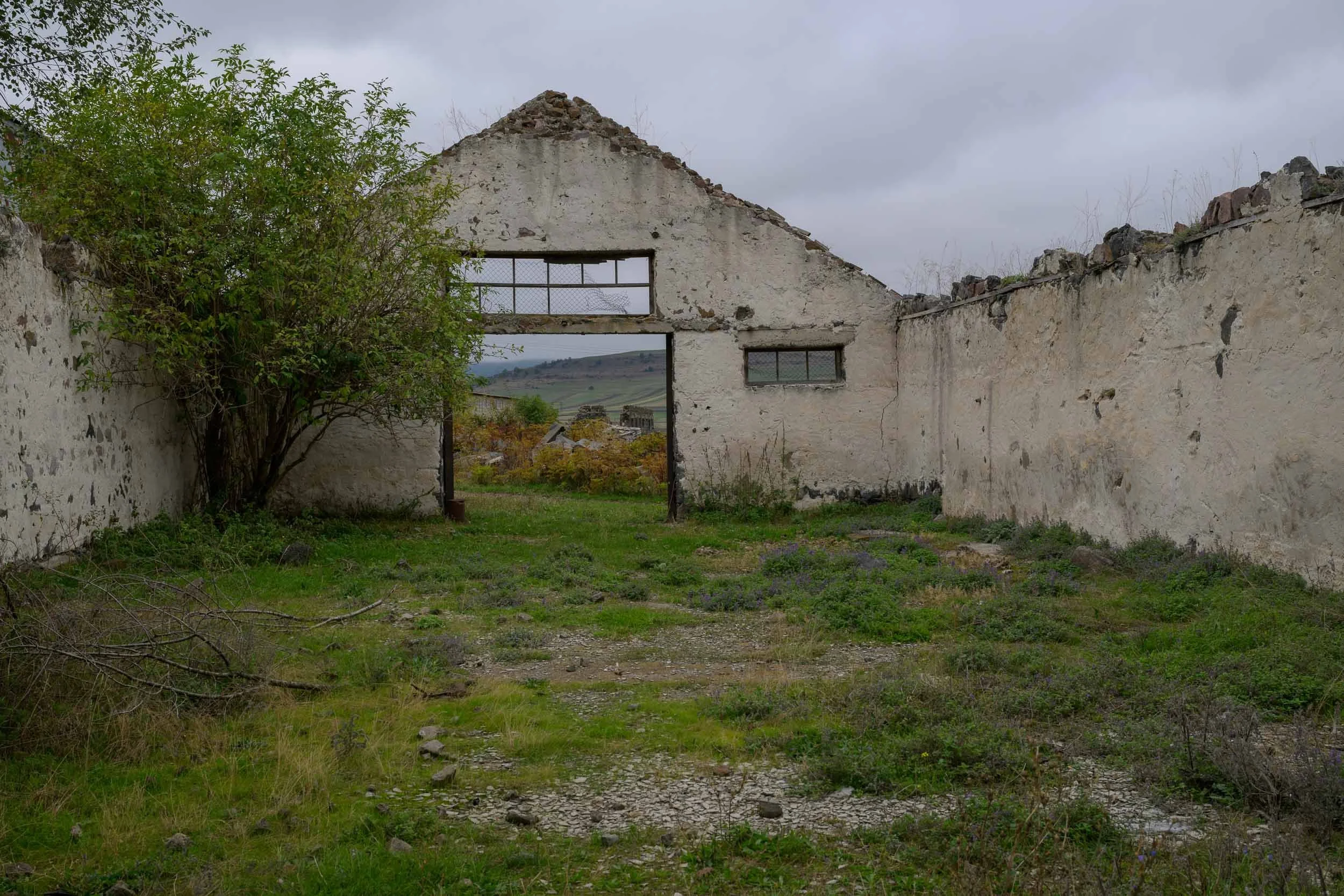 Ruins of an old building with a large open doorway and two small windows, overgrown with grass, shrubs, and small trees, with cloudy sky in the background.