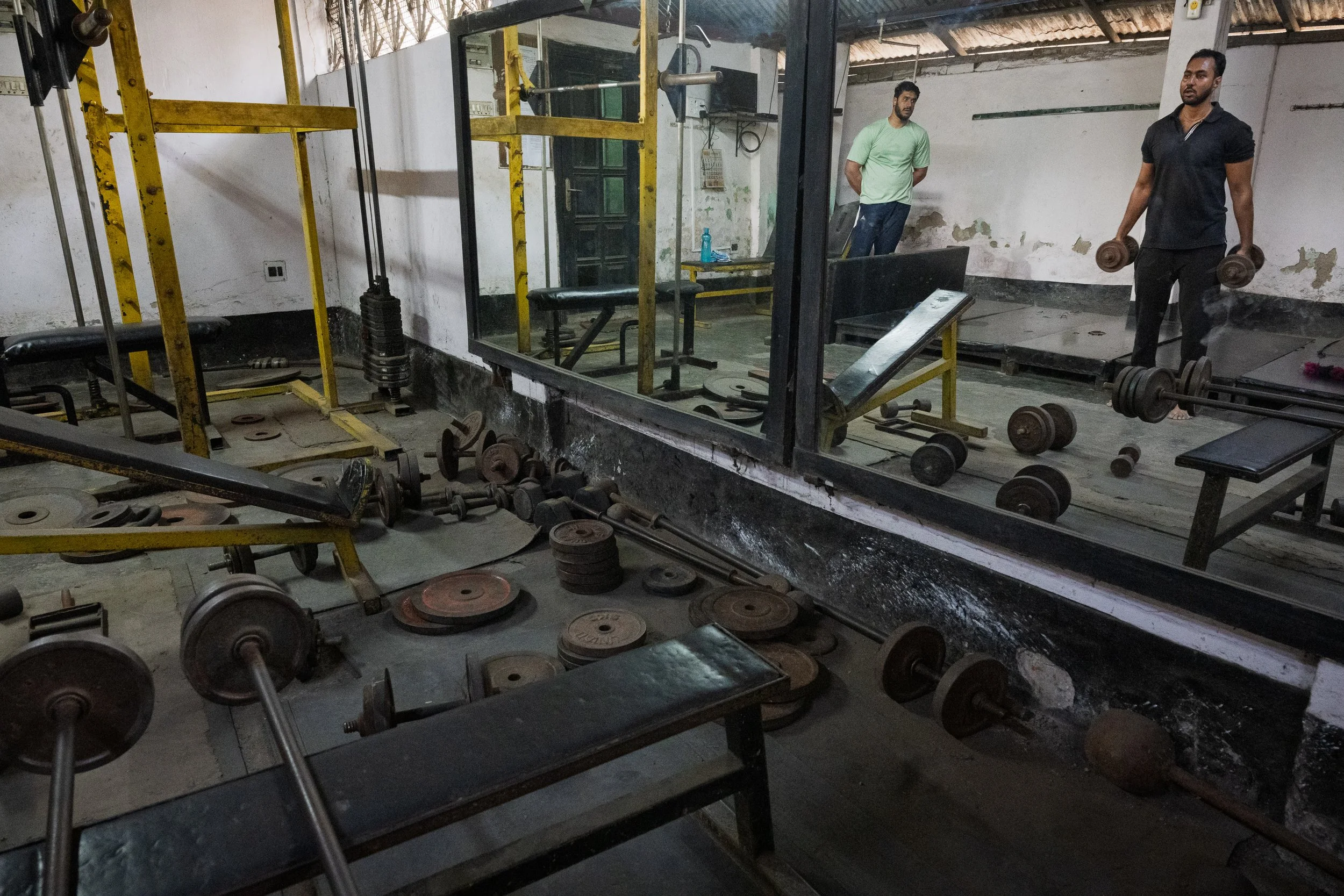 Two men working out with dumbbells in a gym, with various weights scattered around on the floor and mirrors reflecting their image.