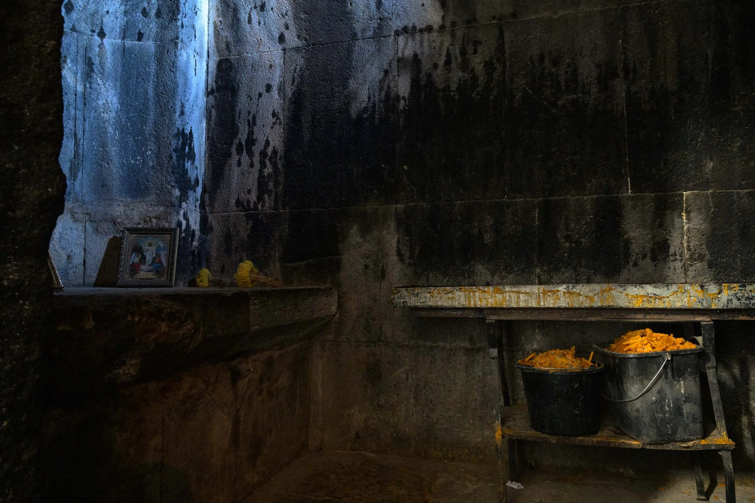 Interior of a small religious shrine or altar with dark stone walls, featuring a framed religious picture, flowers, and offerings, with buckets of yellow flowers and a carved stone or marble slab.