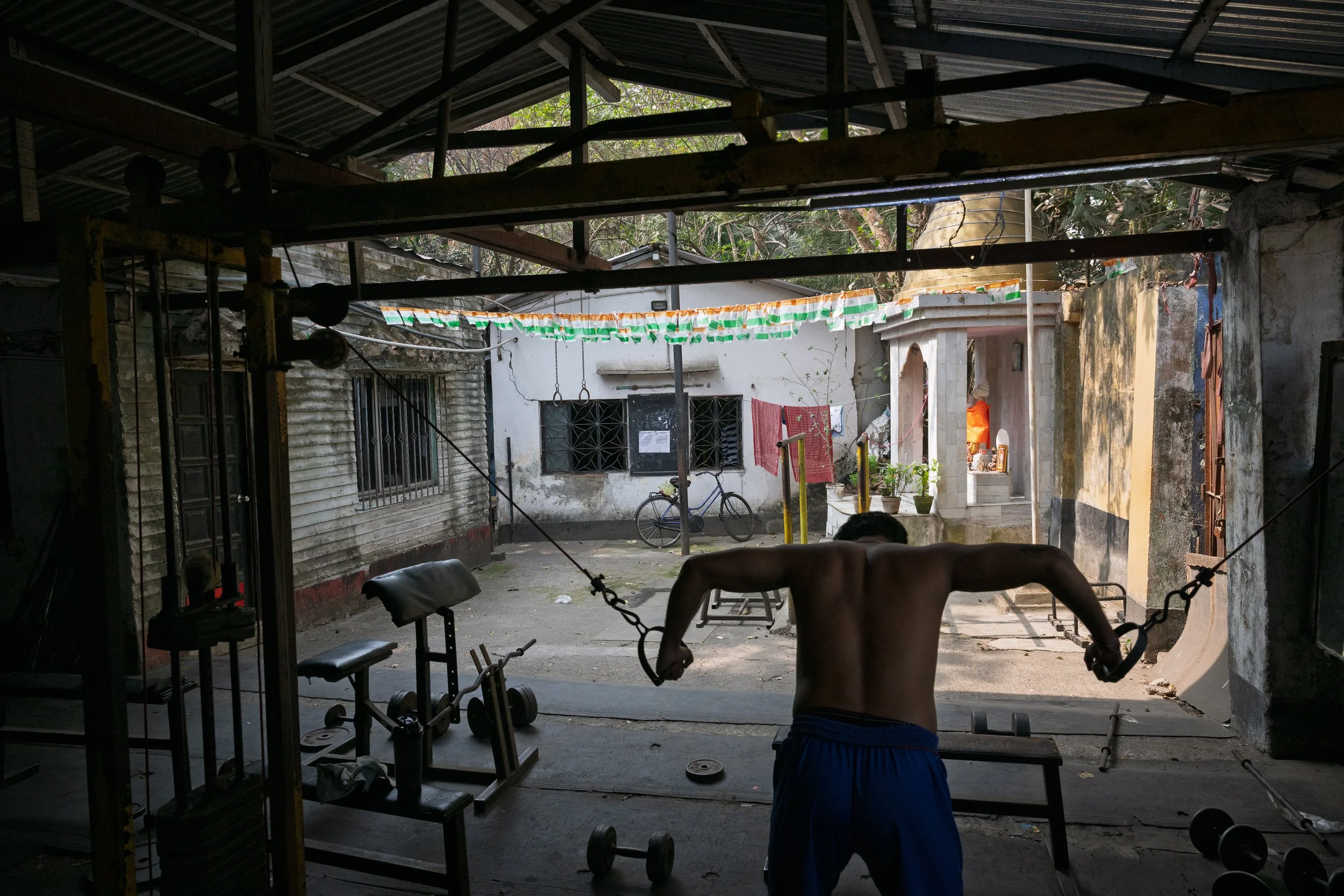 A person with a bare back exercising with resistance bands in an outdoor gym area. The background includes outdoor equipment, a bicycle, laundry hanging, and a small shrine or altar.