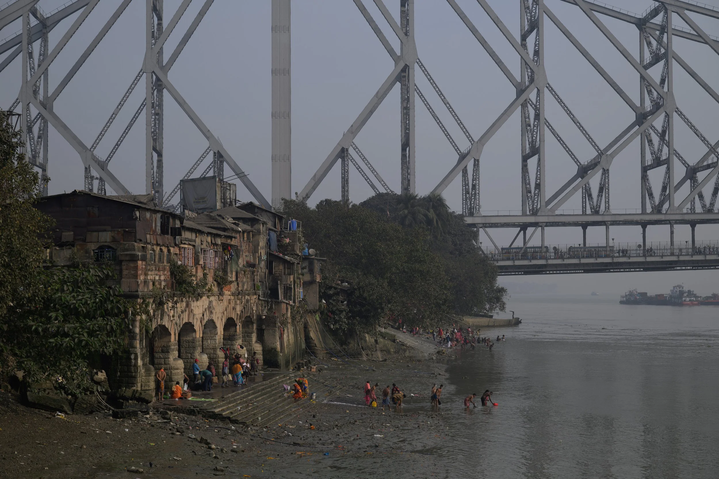 People swimming and washing clothes along a riverbank beneath a large steel bridge, with old buildings lining the shore and cargo ships in the water.