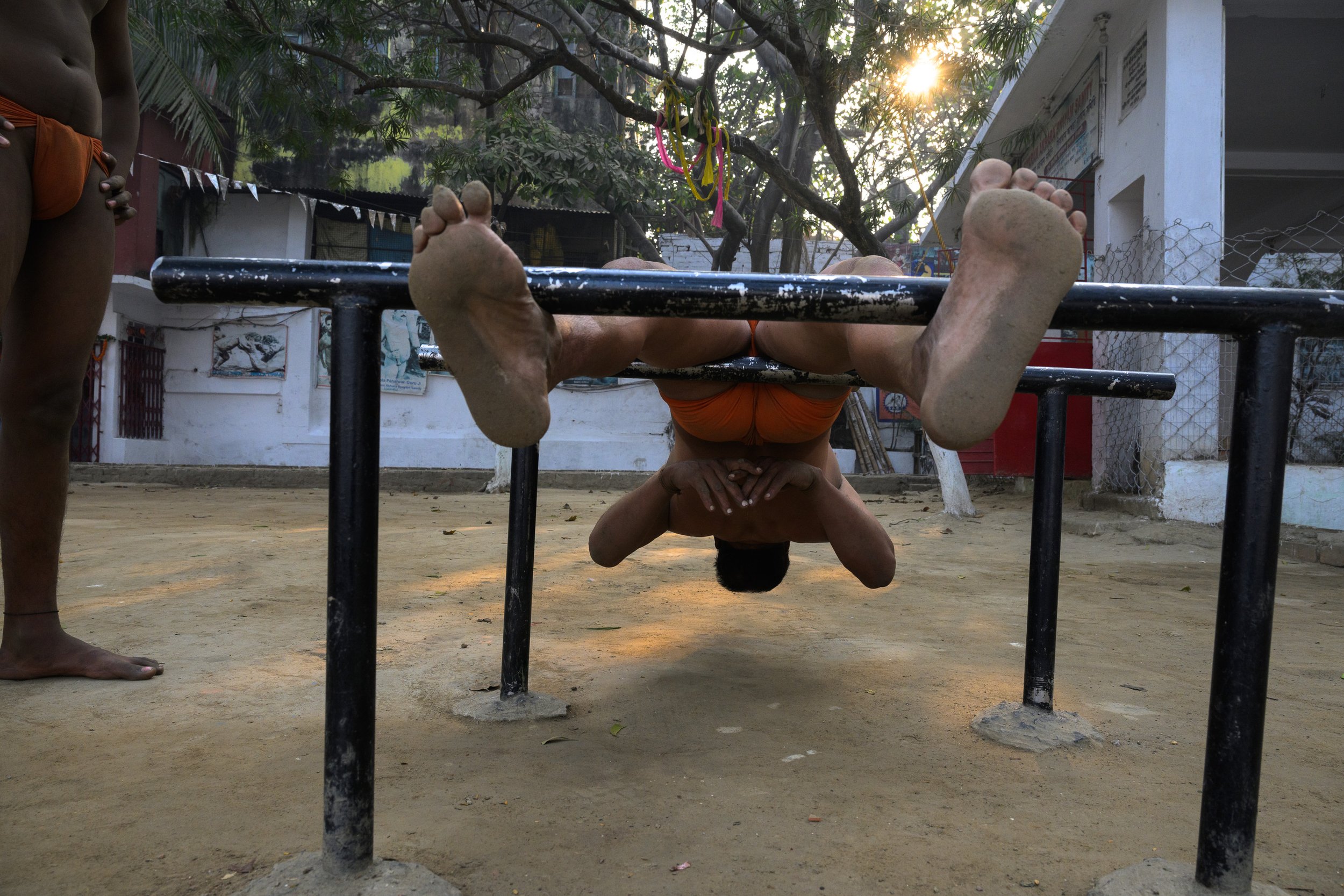 A person performs a headstand on a horizontal bar in an outdoor area, with another person watching nearby. The person is upside down with legs spread wide apart and hands clasped together at their neck. The scene is set in a dirt courtyard with trees