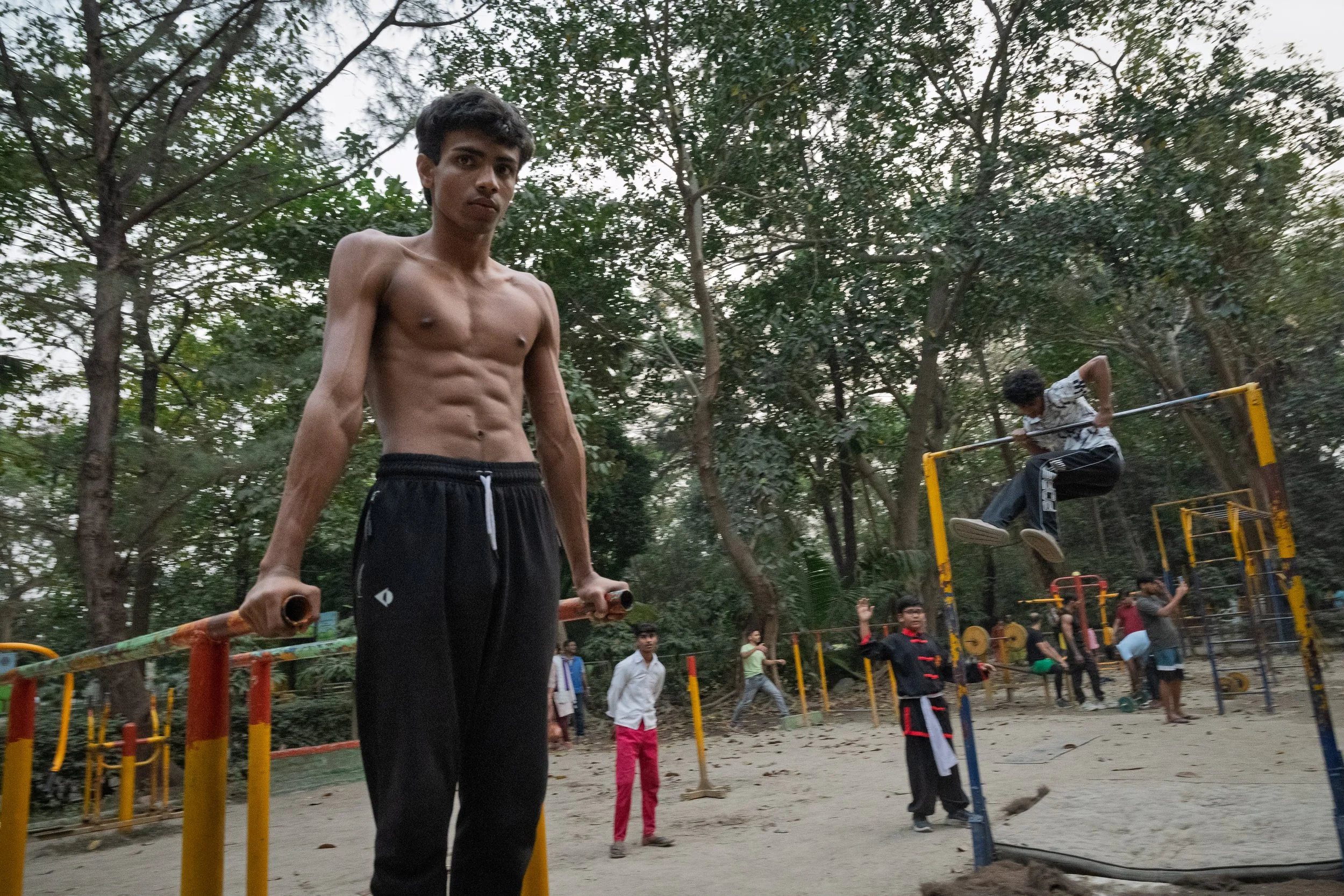 A shirtless young man with black pants stands holding parallel bars, with children and adults in the background at an outdoor exercise area surrounded by trees. One child is jumping over a horizontal bar, while others watch or engage in different act