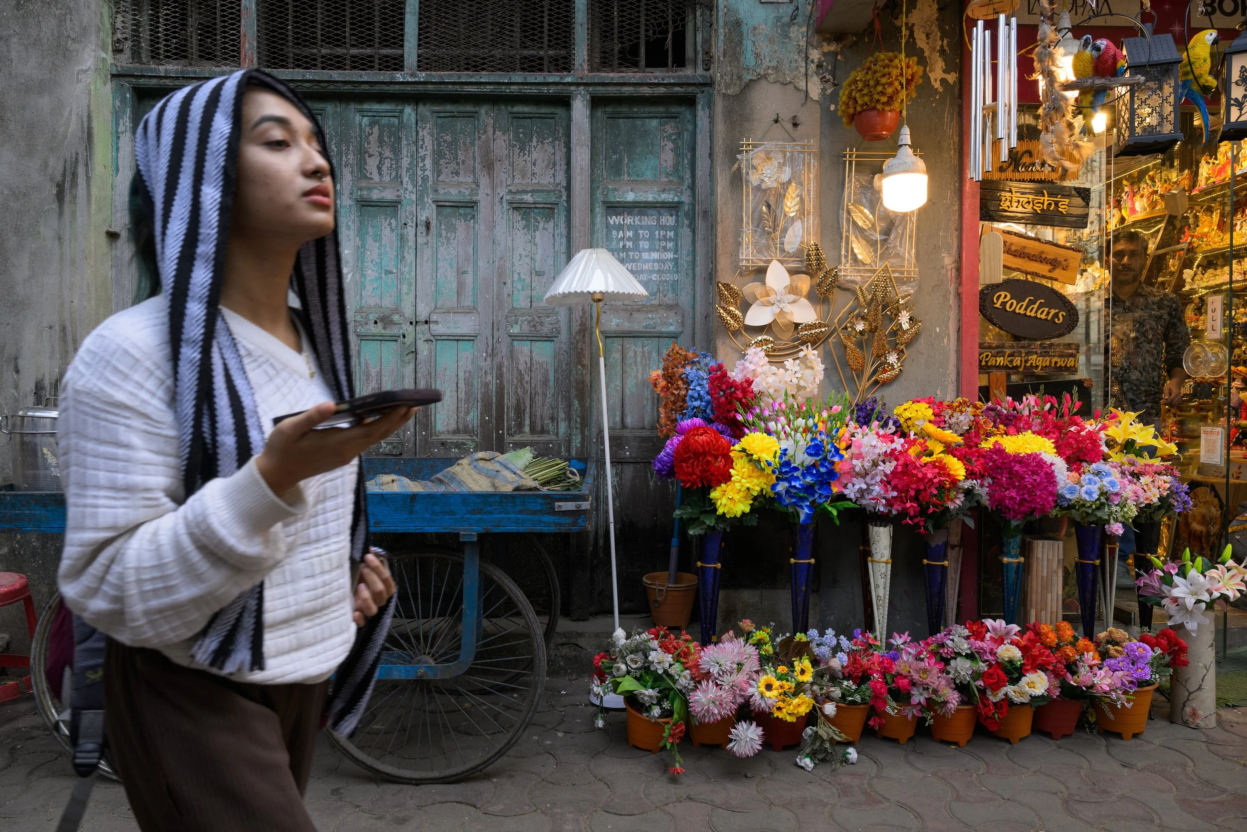 A young woman walking past a flower shop with colorful flower bouquets outside on the street in an outdoor market setting.