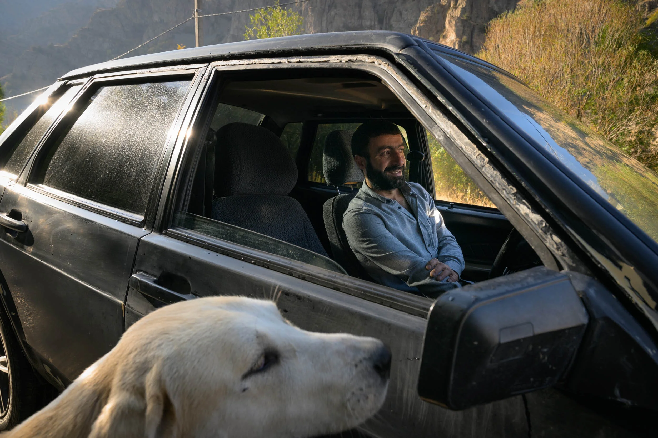 A man with a beard sitting inside a black car, smiling, with a backdrop of trees and hills. A dog’s nose and part of its face are visible in the foreground, near the open driver’s side window.
