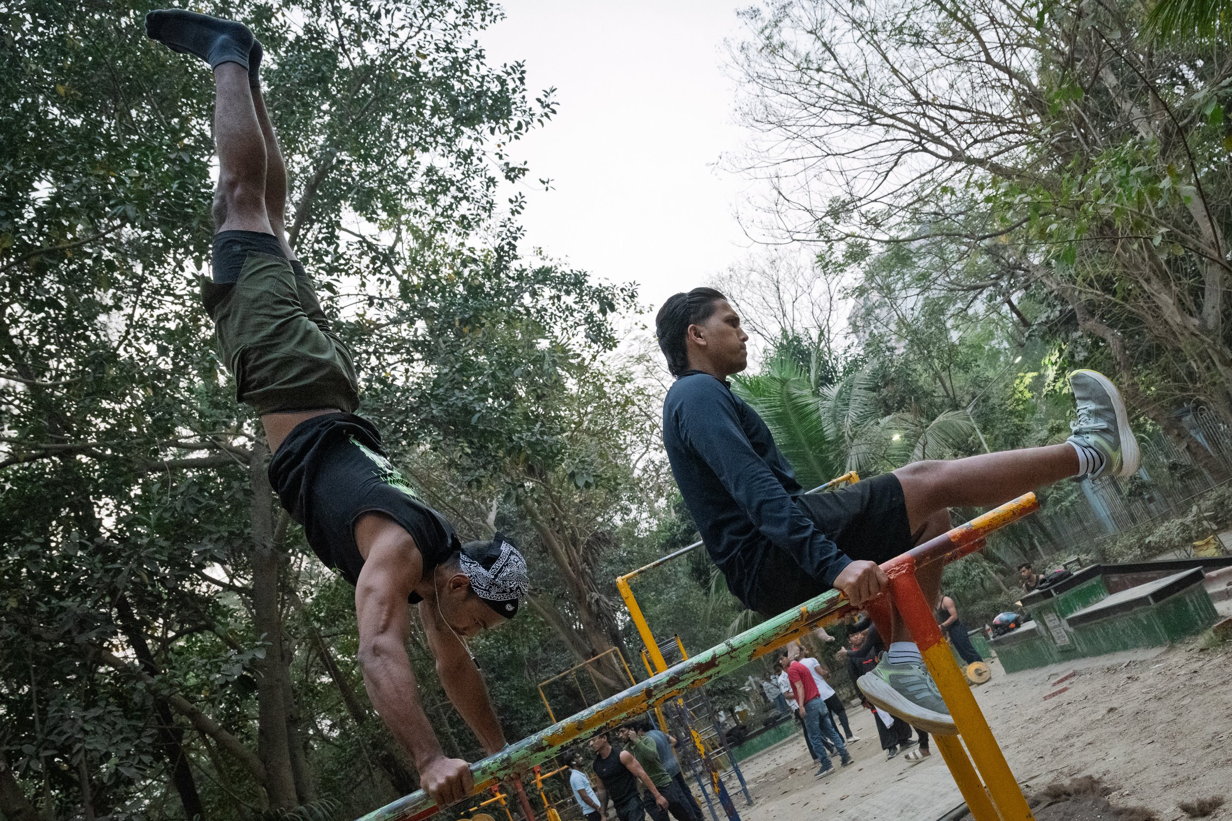 Two young men are performing acrobatic yoga poses on a jungle gym in a park, with trees and people in the background.