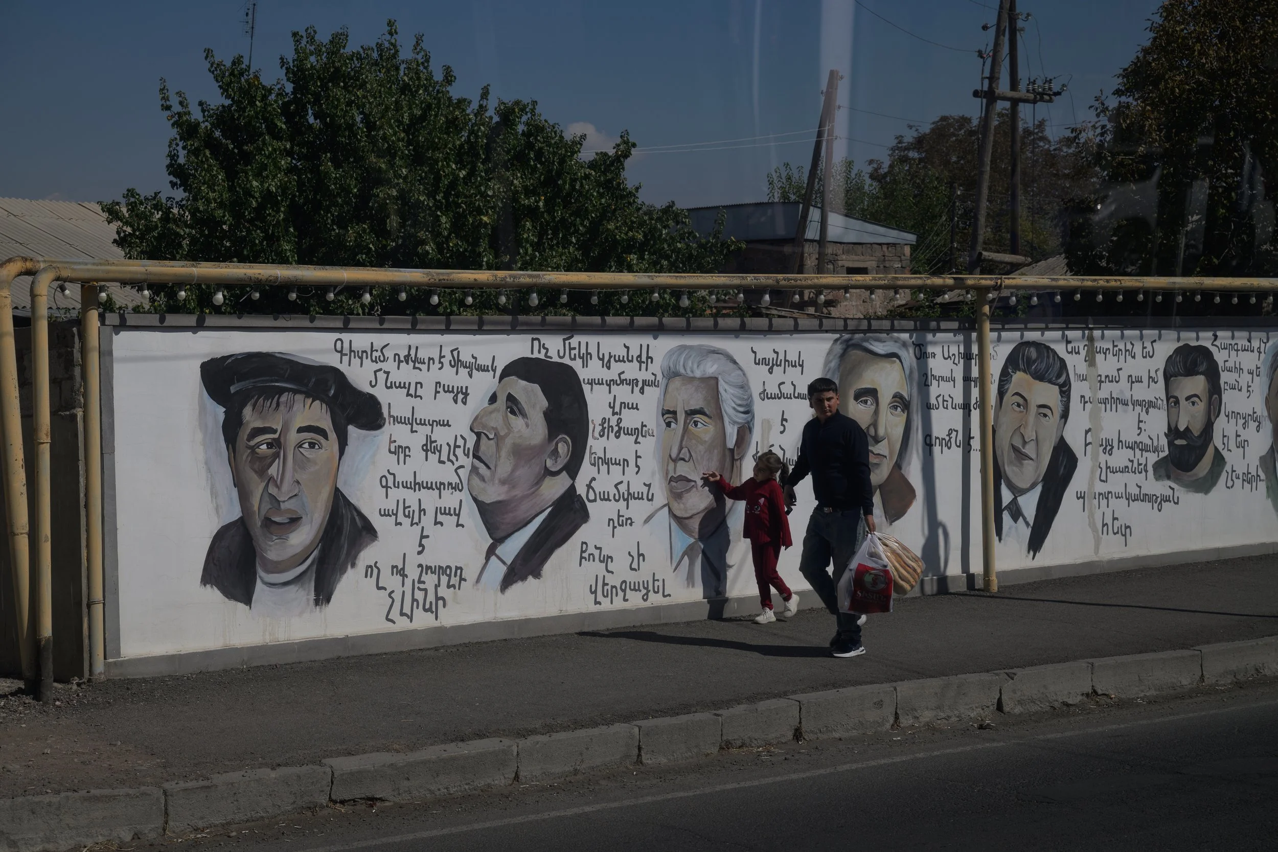 Mural of multiple illustrated faces of men with writing in Armenian script on a white wall. People walking past in front of the mural.