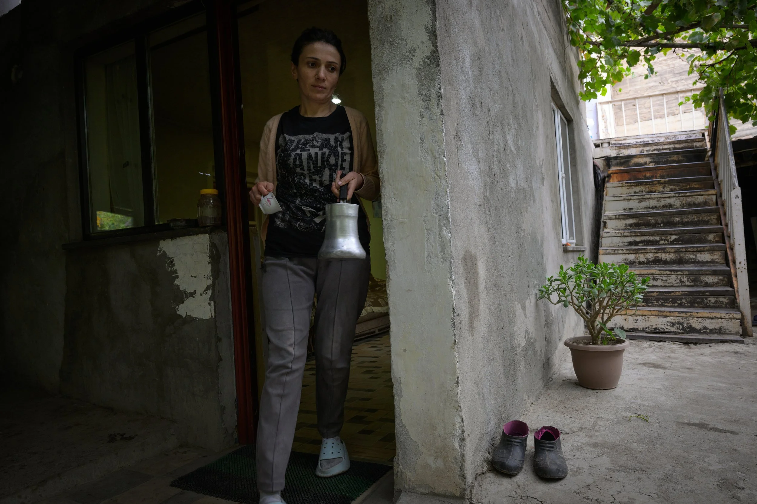 A woman with dark hair stands at the entrance of a house holding a kettle and a small container. She wears a black graphic T-shirt, gray pants, and white Crocs. Outside, there is a potted plant and a pair of black shoes on the ground near a staircase