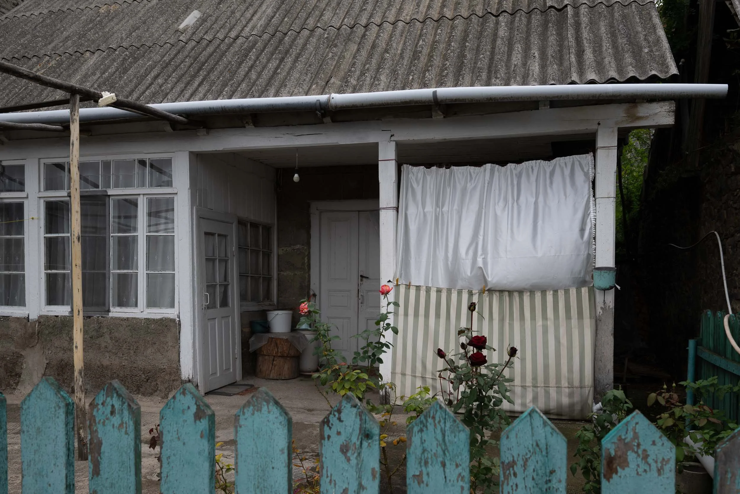 The exterior of a house with a corrugated roof, a white door, and a curtain covering part of the porch. There is a wooden fence painted blue, some pink roses, and gardening tools in the yard.