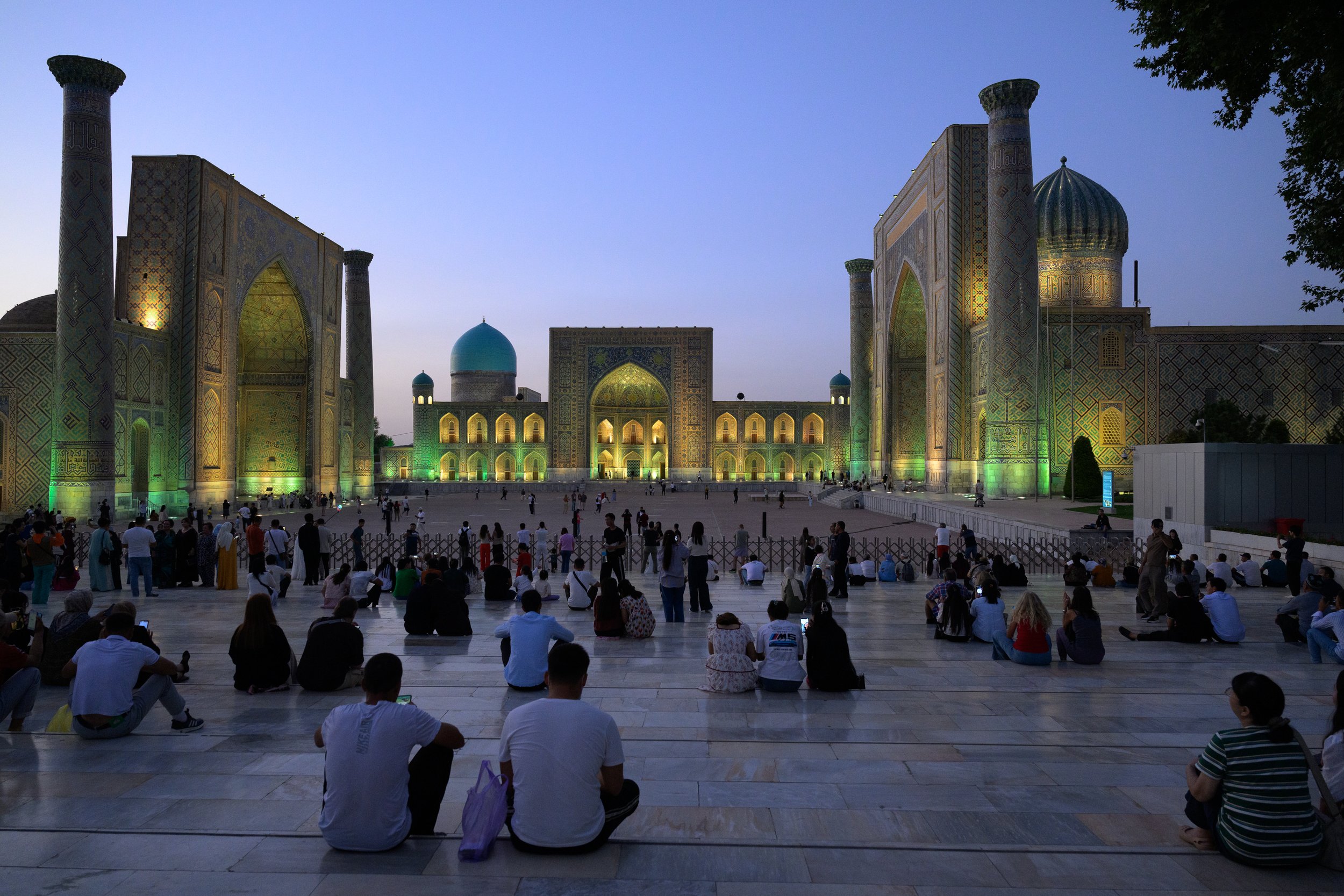 Large mosque illuminated at dusk, with many people sitting and standing in front of it, some taking pictures.