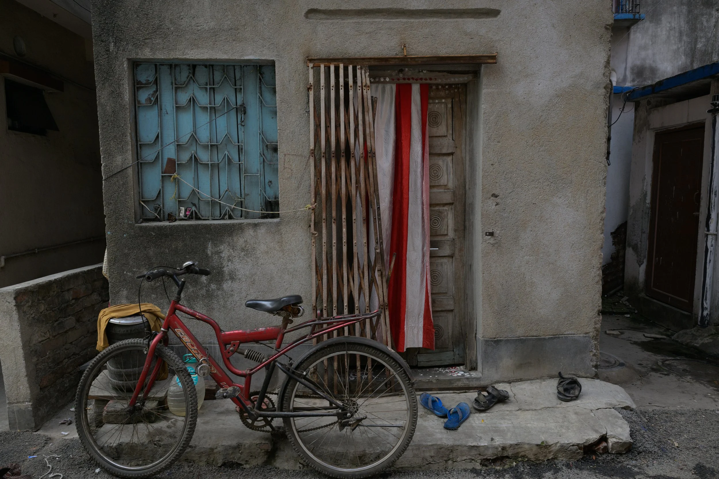 A red bicycle parked outside a weathered concrete house with a barred window and a partly covered door with a red and white curtain. There are three pairs of shoes on the ground, and a small container next to the bicycle.