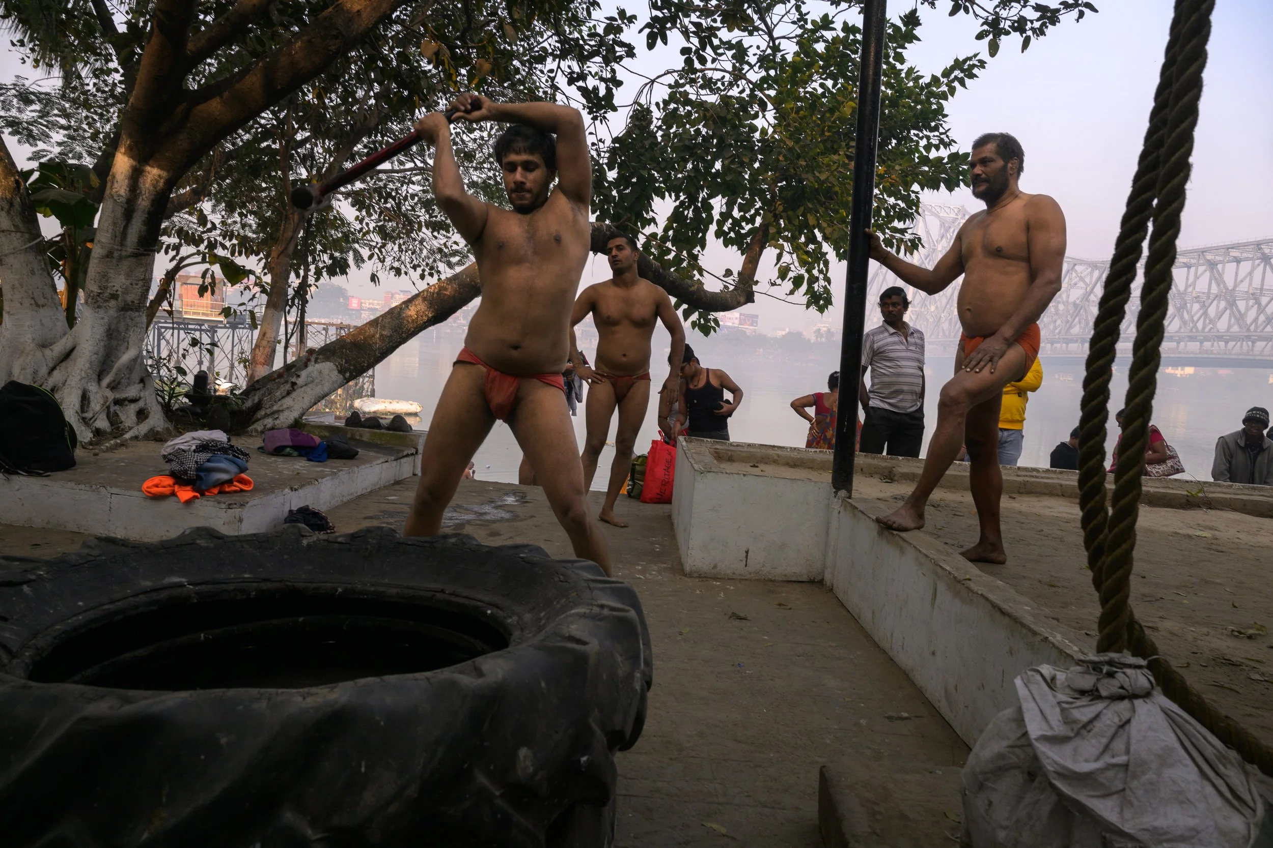 Men in loincloths exercising outdoors near a large tree and a body of water, with onlookers in the background.