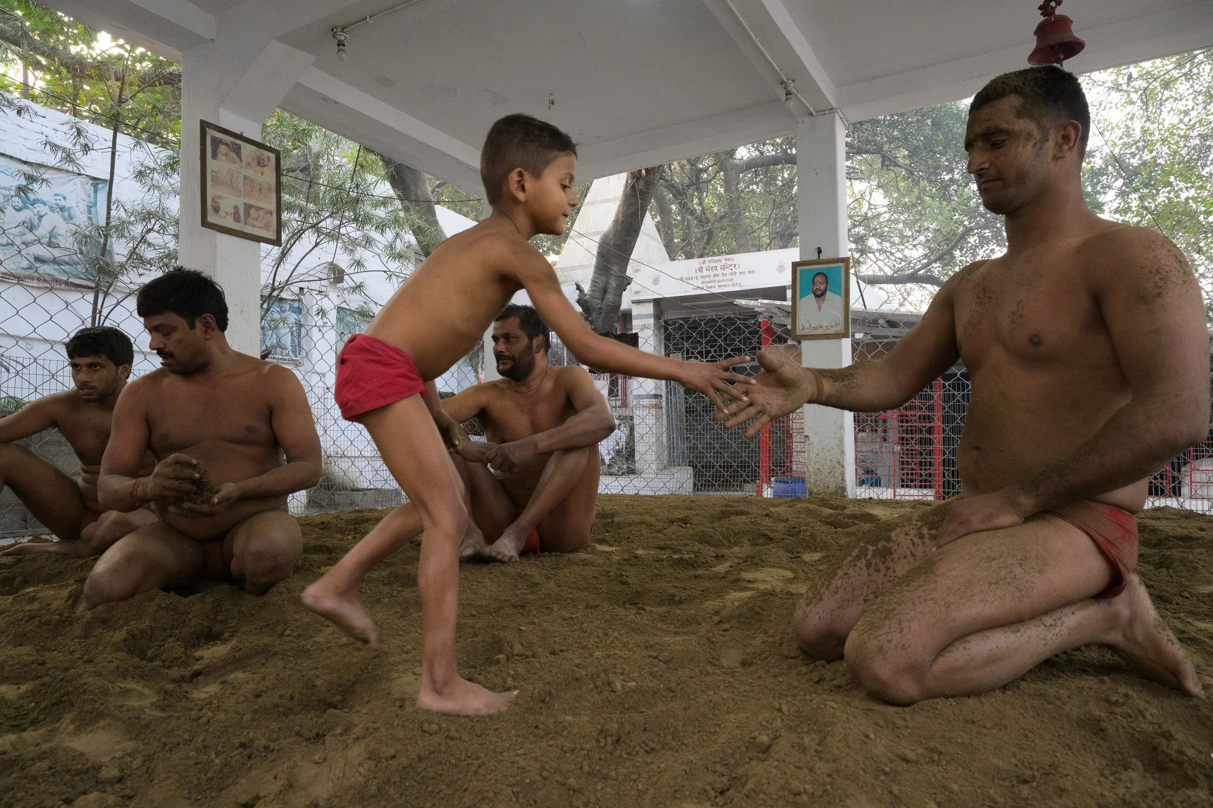 A young boy in red shorts is reaching out to shake hands with a man kneeling on sandy ground, who is also shirtless and wearing red shorts. Several other shirtless men are nearby, sitting on the sand, with trees and a white fence in the background.