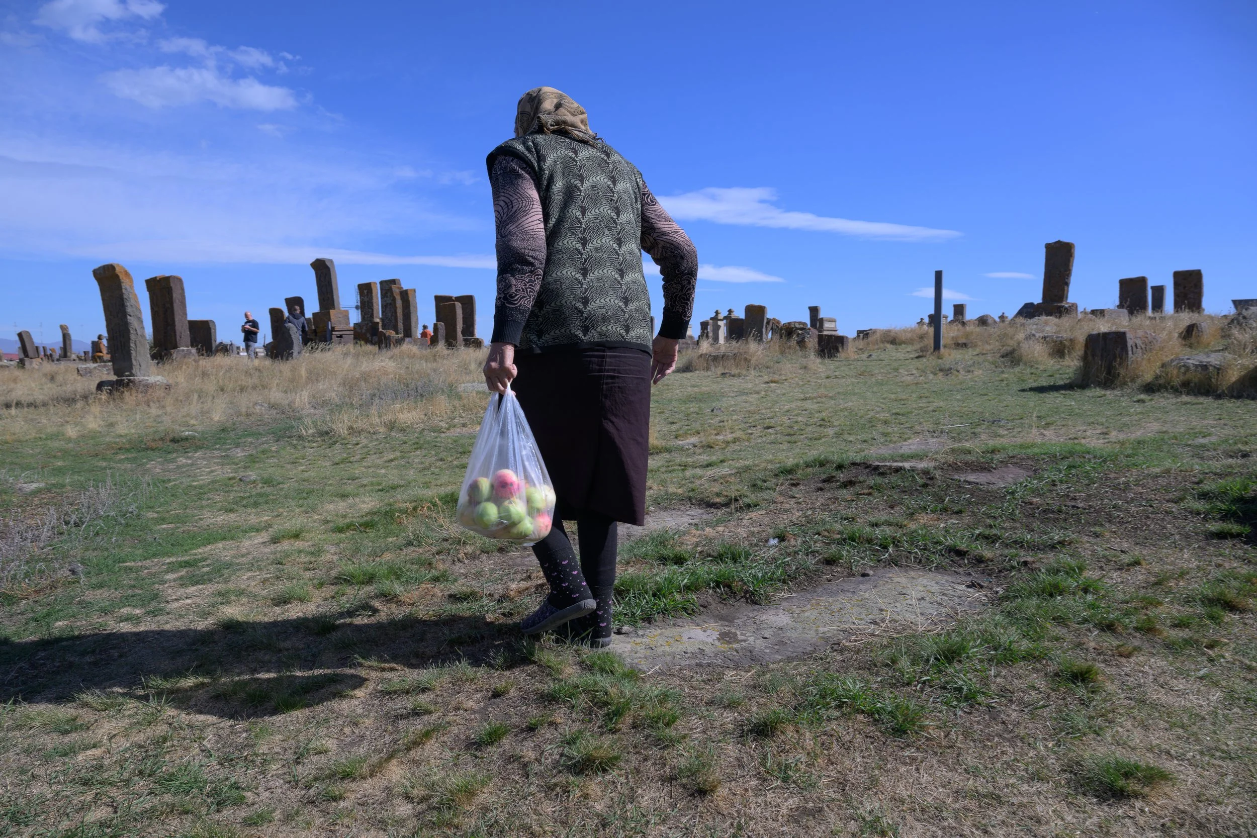 A woman walking on grass with a plastic bag full of apples, in an open field with ancient stone ruins under a blue sky.
