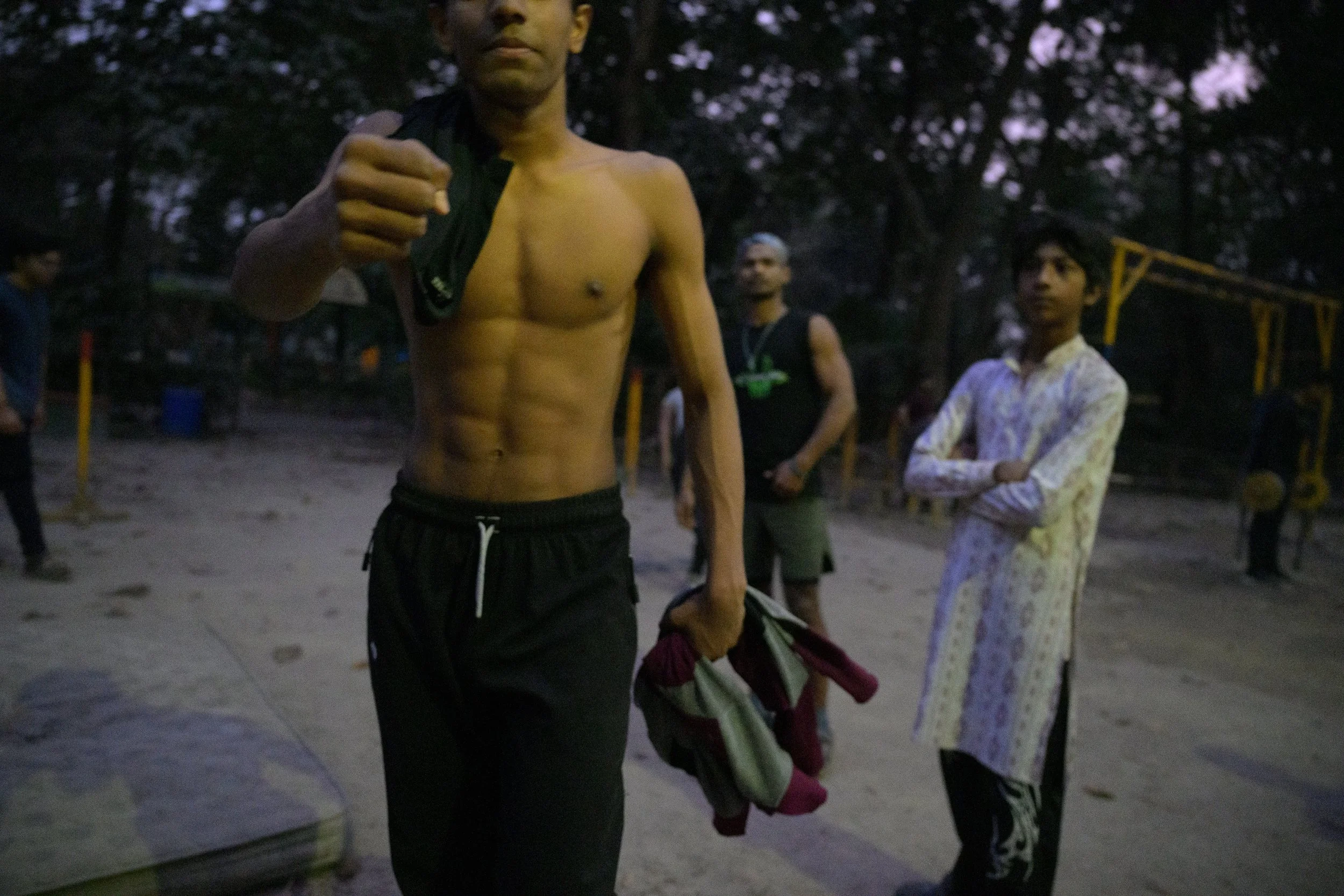 A shirtless man with a towel over his shoulder giving a thumbs-up, standing outdoors at dusk with several people in the background.