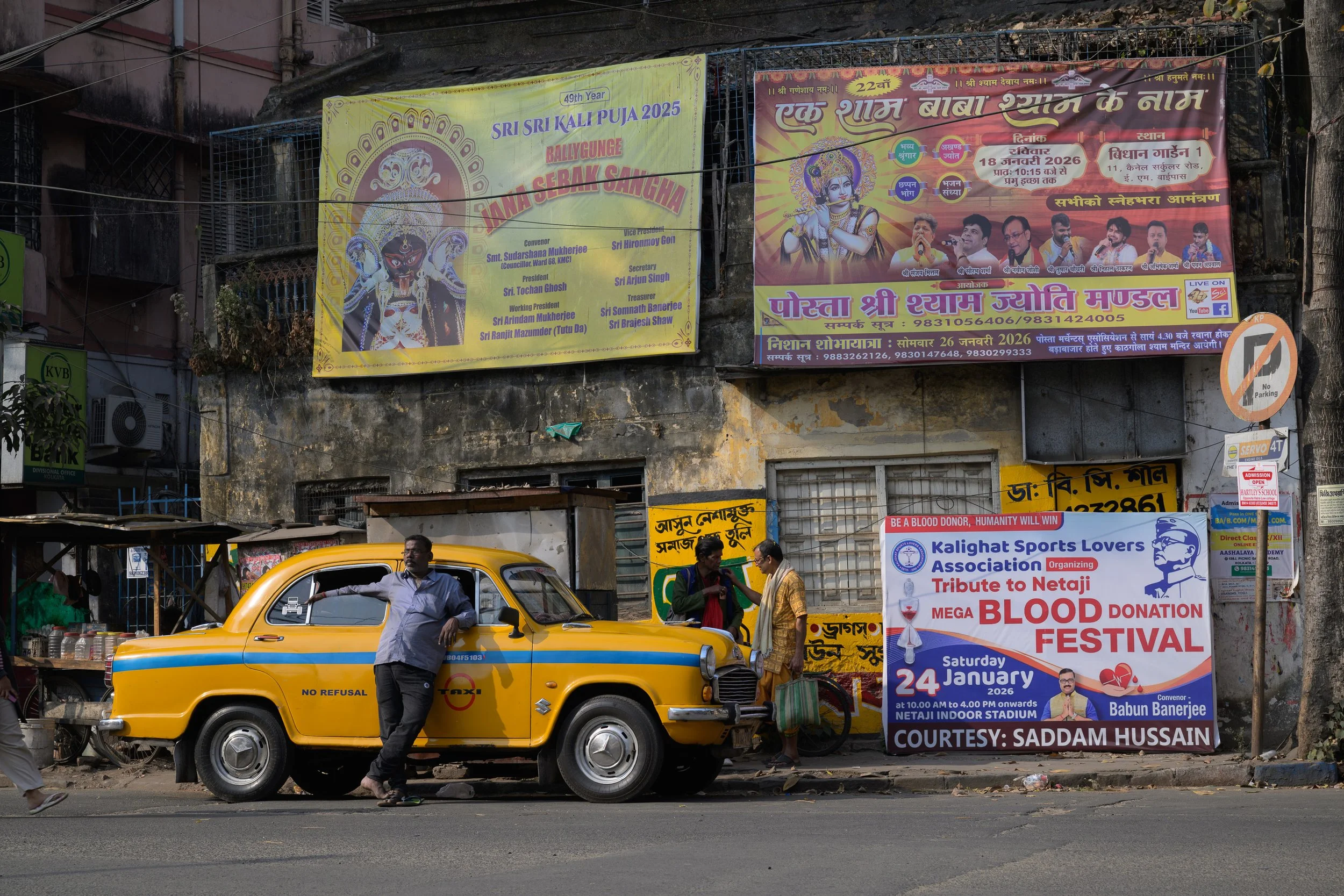 A man leaning against a yellow taxi parked on the street, with two women standing nearby and engaged in conversation, behind a small street vendor stall, with various colorful posters and signs on the wall behind them, including an advertisement for 