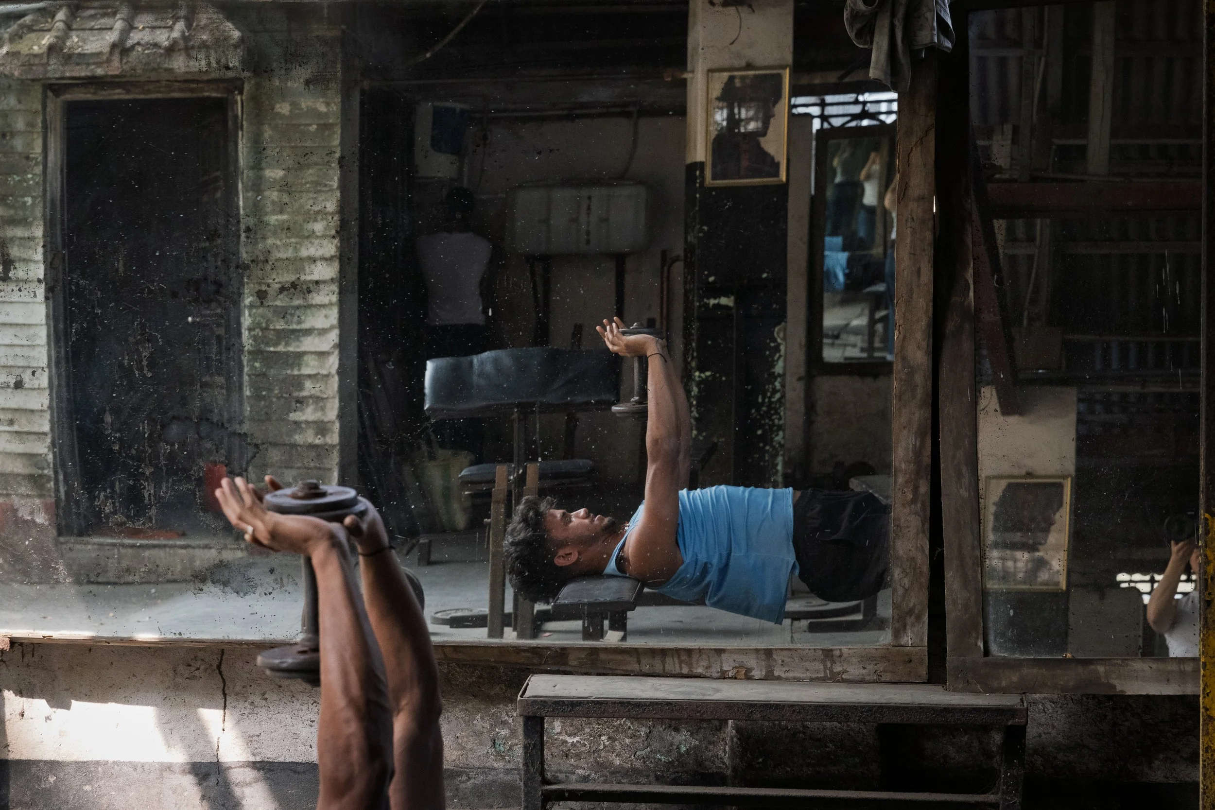 A person lying on a gym bench in a gym, seen through a mirror, with their arm extended and holding a dumbbell, in a rustic gym with exposed brick walls and gym equipment.