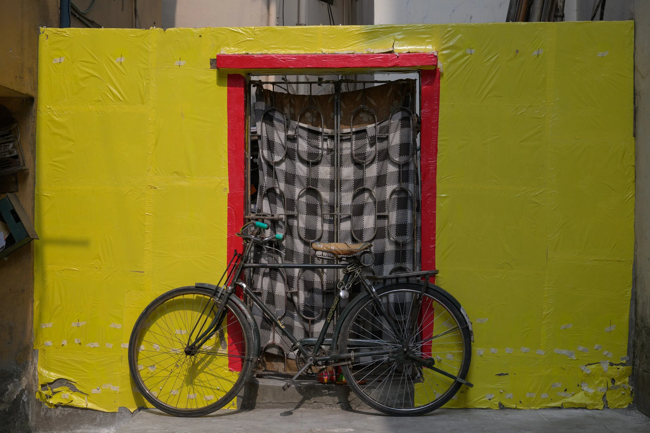 A black bicycle with a tan leather seat leaning against a yellow wall with red trim and a decorative iron gate in the center.
