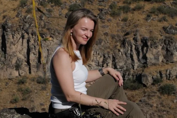 A woman with long blonde hair wearing a white tank top and brown pants sitting outdoors on a rocky hillside.