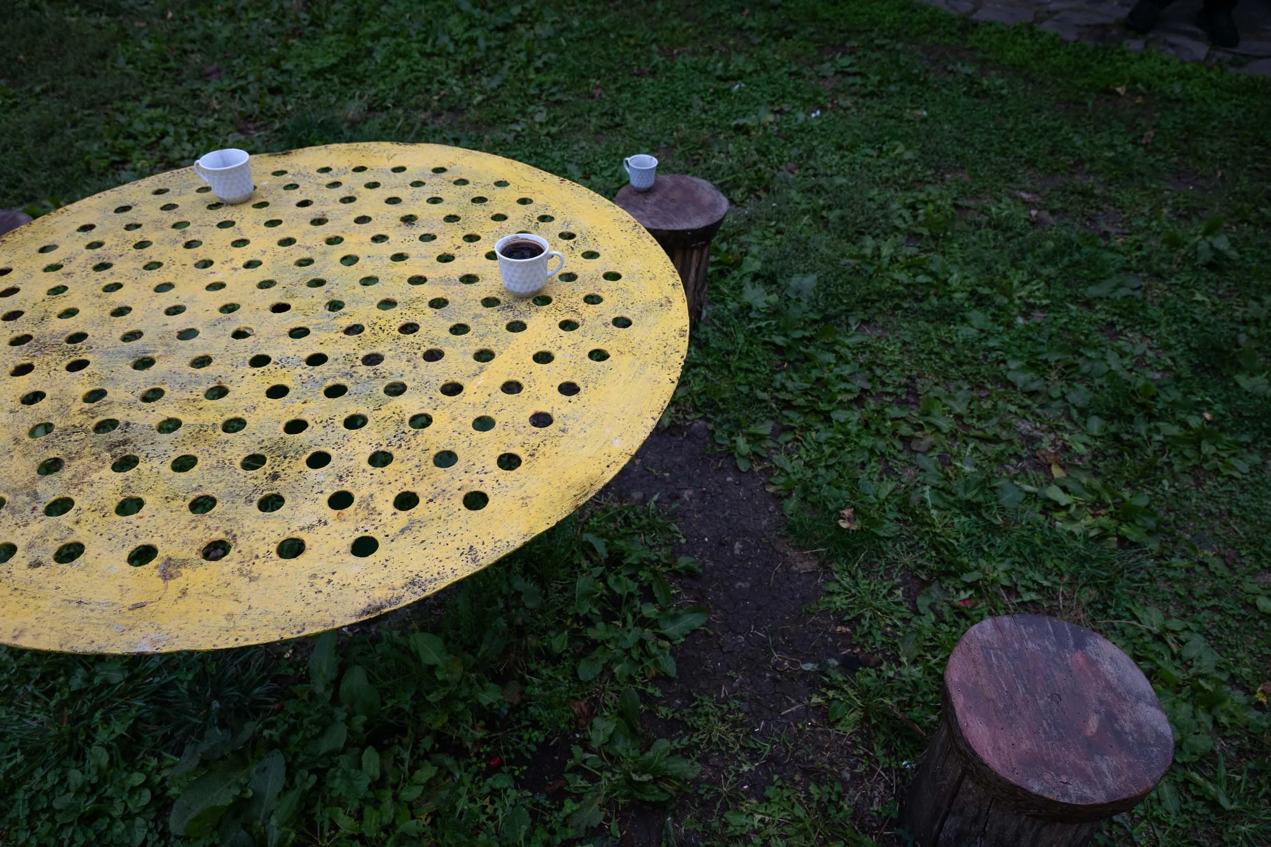 Yellow perforated metal table with three white coffee mugs, one filled with coffee, in an outdoor grassy area with tree stumps.