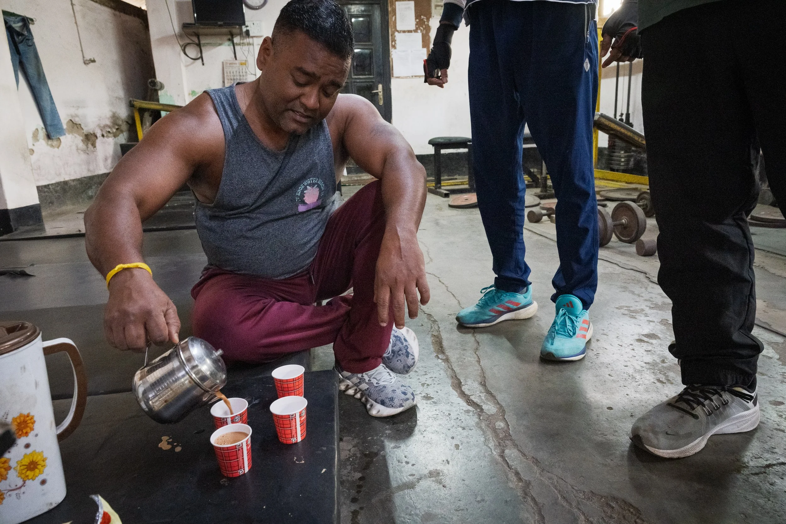 A man is sitting on the floor pouring tea from a metal pot into red checkered paper cups, with three cups arranged in a row.