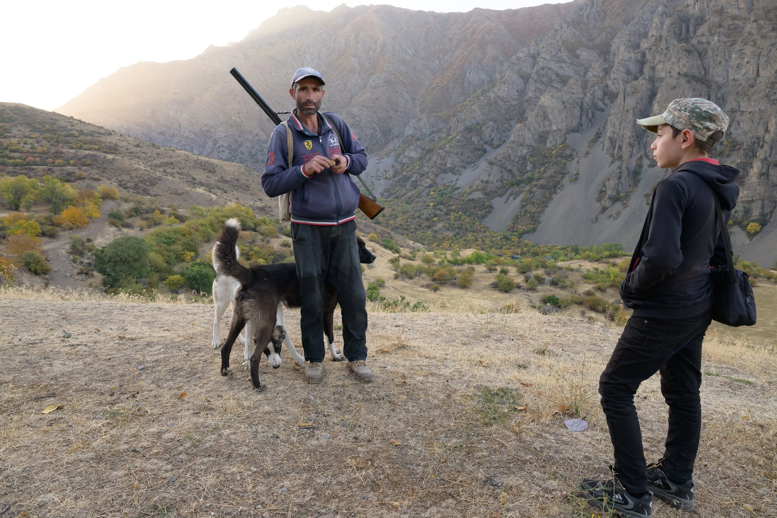 A man and a boy stand outdoors in a mountainous landscape, accompanied by a dog. The man holds a rifle, and they appear to be in a conversation or preparing for an activity. The scene is during daytime with autumn foliage on the hillsides.