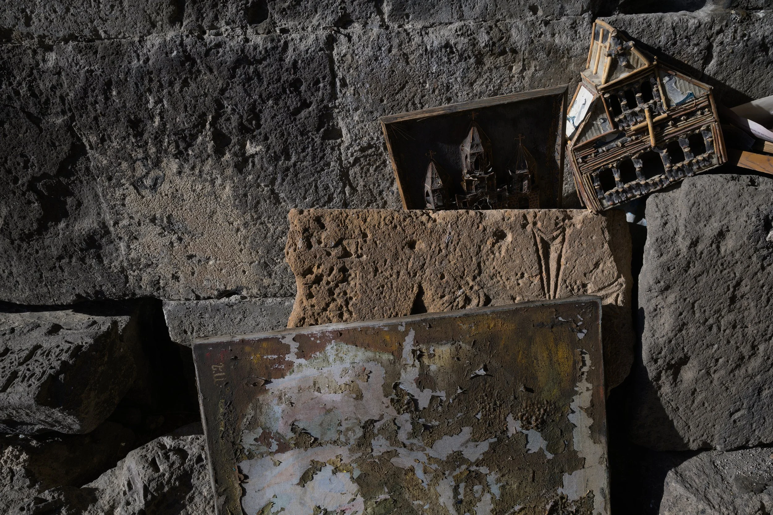 A collection of old, weathered objects including a rusty metal sheet, small wooden carved houses, and a stone with carved crosses, all resting against large rough stones.