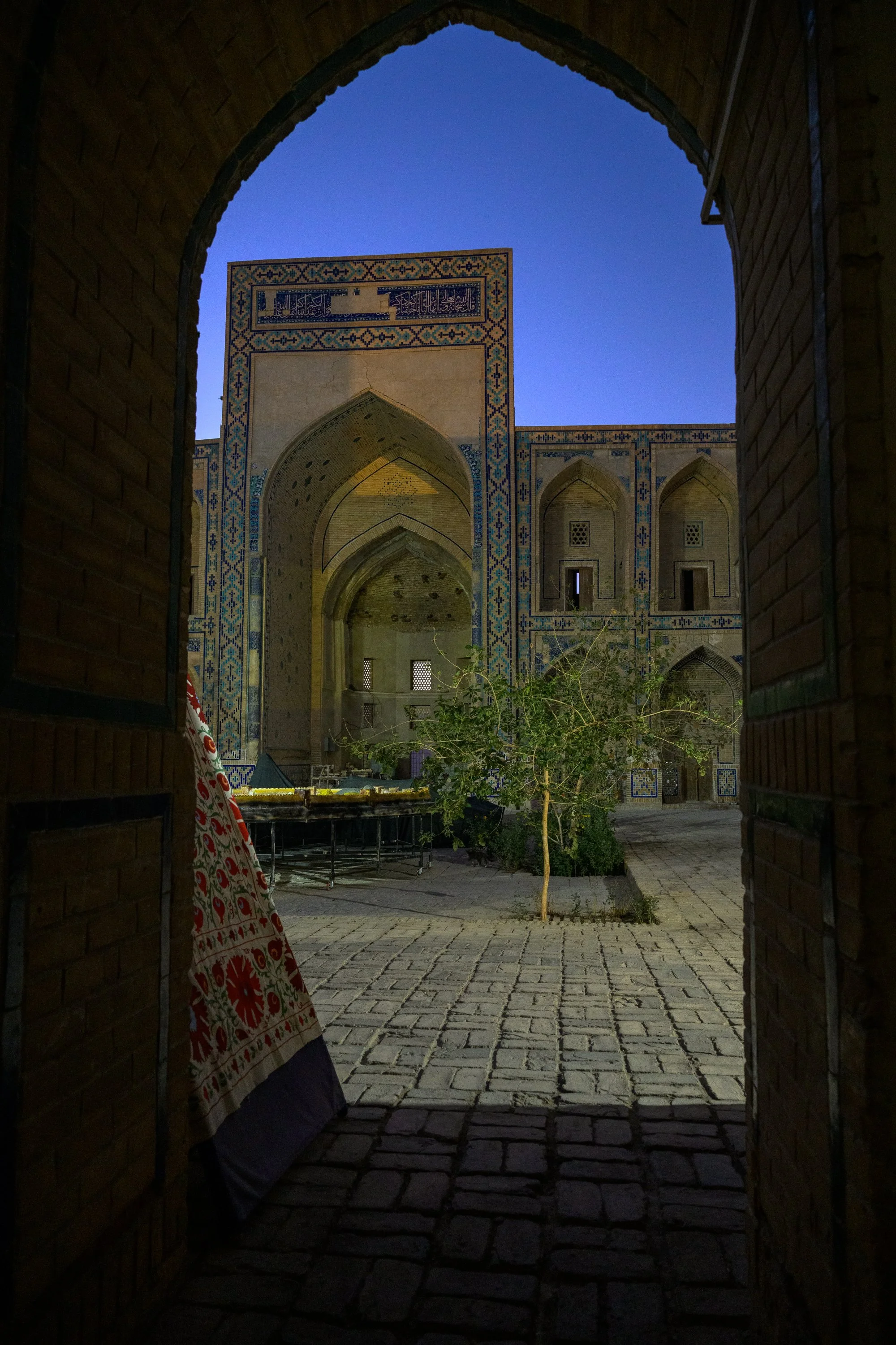 View through a brick archway of a historic building with decorative tile work, arched windows, and a courtyard with cobblestone paving and a small tree, under a twilight sky.