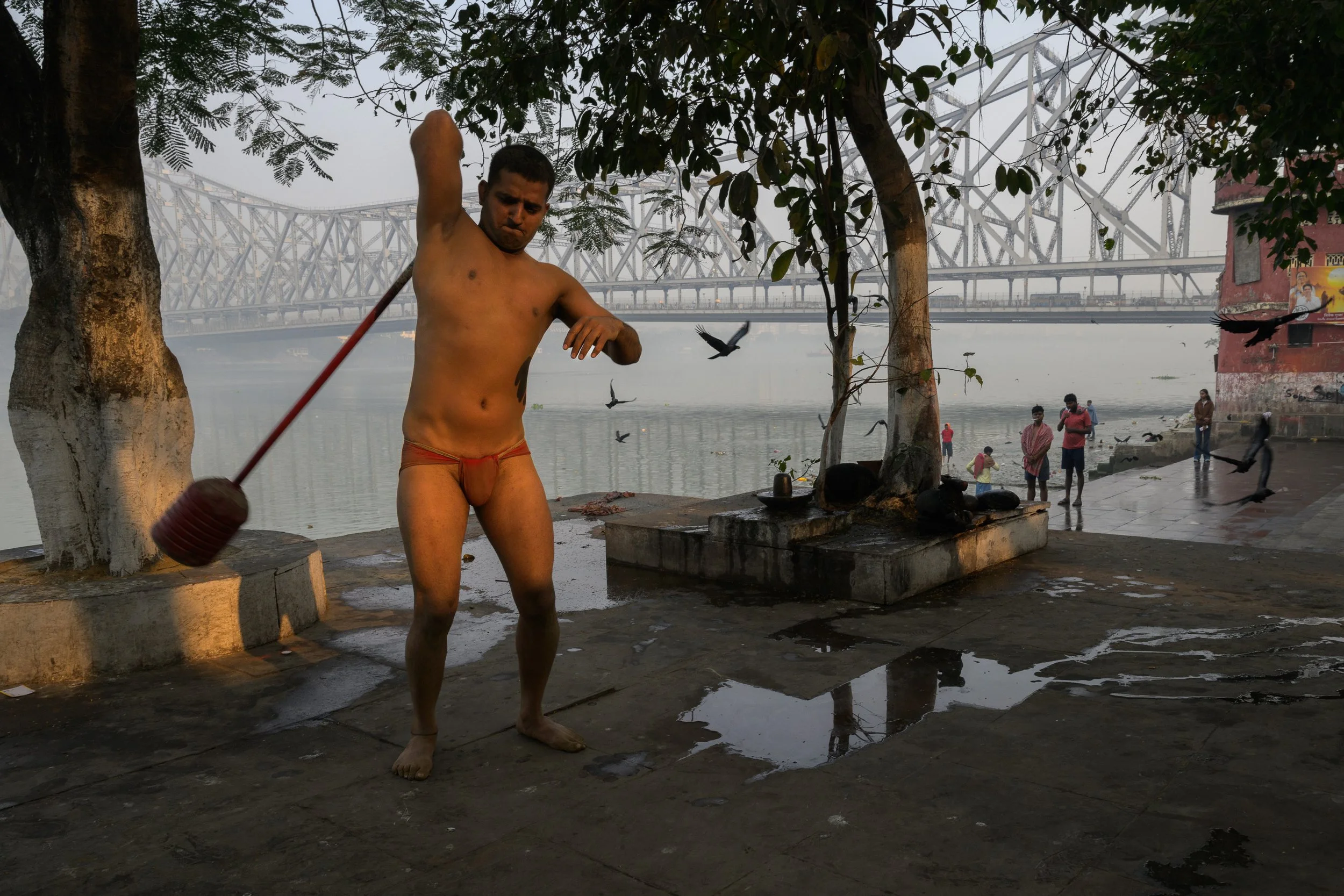 Man standing outdoors near water, wearing only red underwear, holding a traditional wood and rubber hammer, with trees, a bridge, and people in the background.