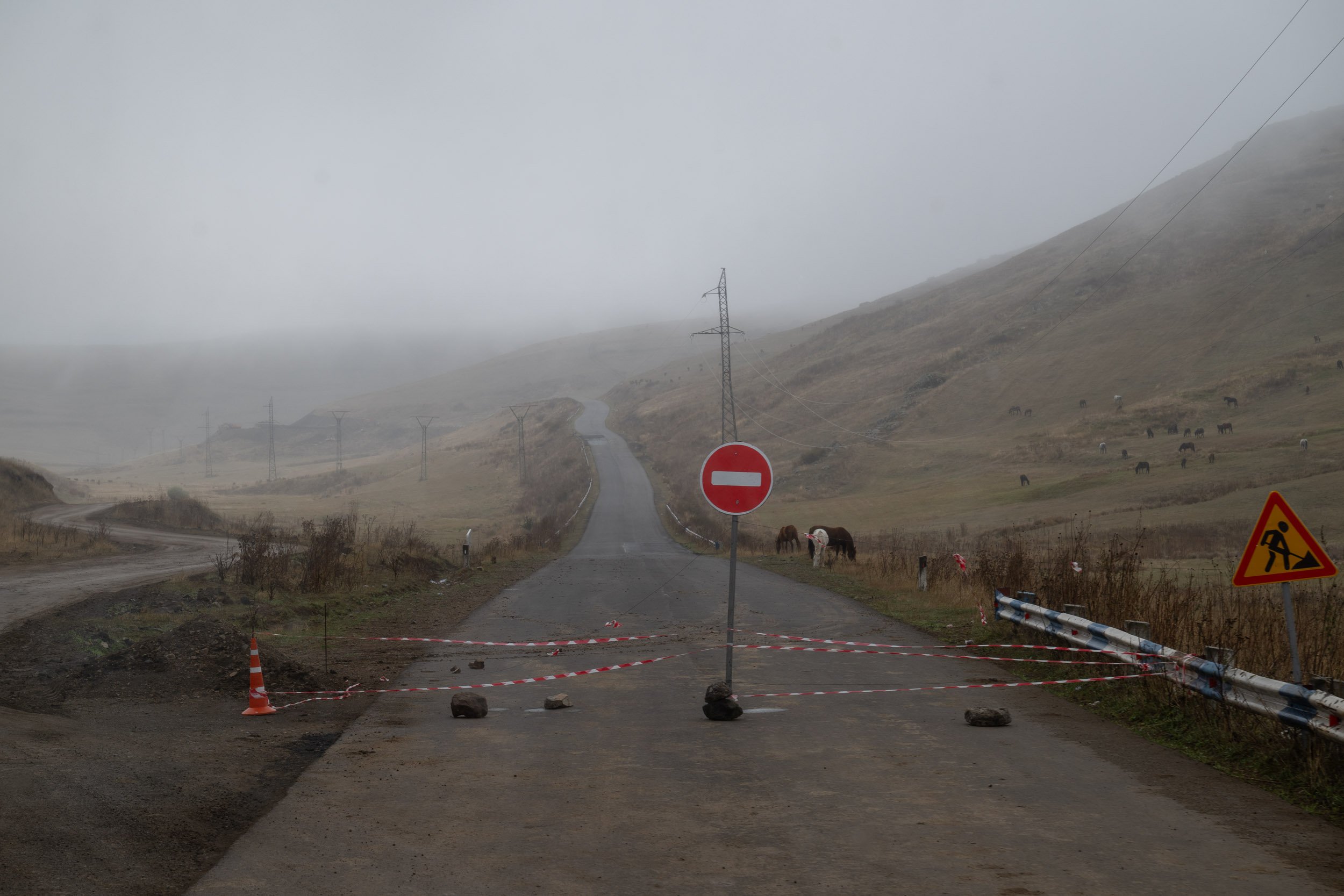 A foggy rural road blocked by construction barriers with a no entry sign, power lines, hills, grazing horses, and construction warning signs visible in the background.