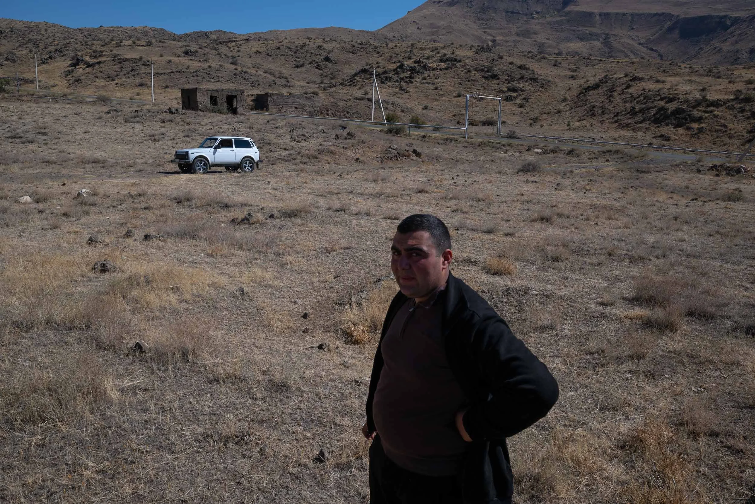A man with dark hair, wearing a black jacket, standing in a dry, grassy, mountainous landscape with a white compact SUV in the background. There are utility poles and an old, abandoned building in the distance.