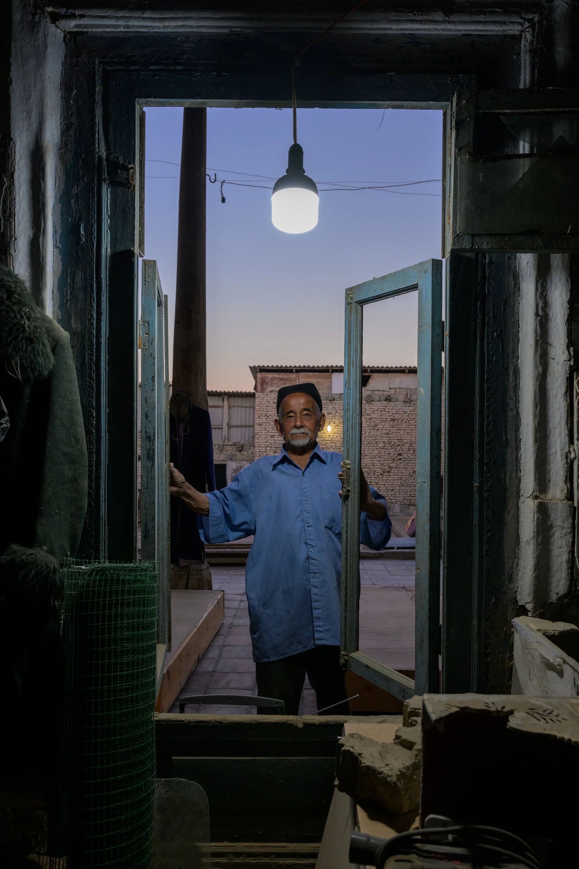 An elderly man with gray hair and beard, wearing a blue shirt, stands at an open door, holding the door frame with both hands, looking at the camera. The sky is in twilight, and the scene is indoors with various objects on the left and a brick wall o
