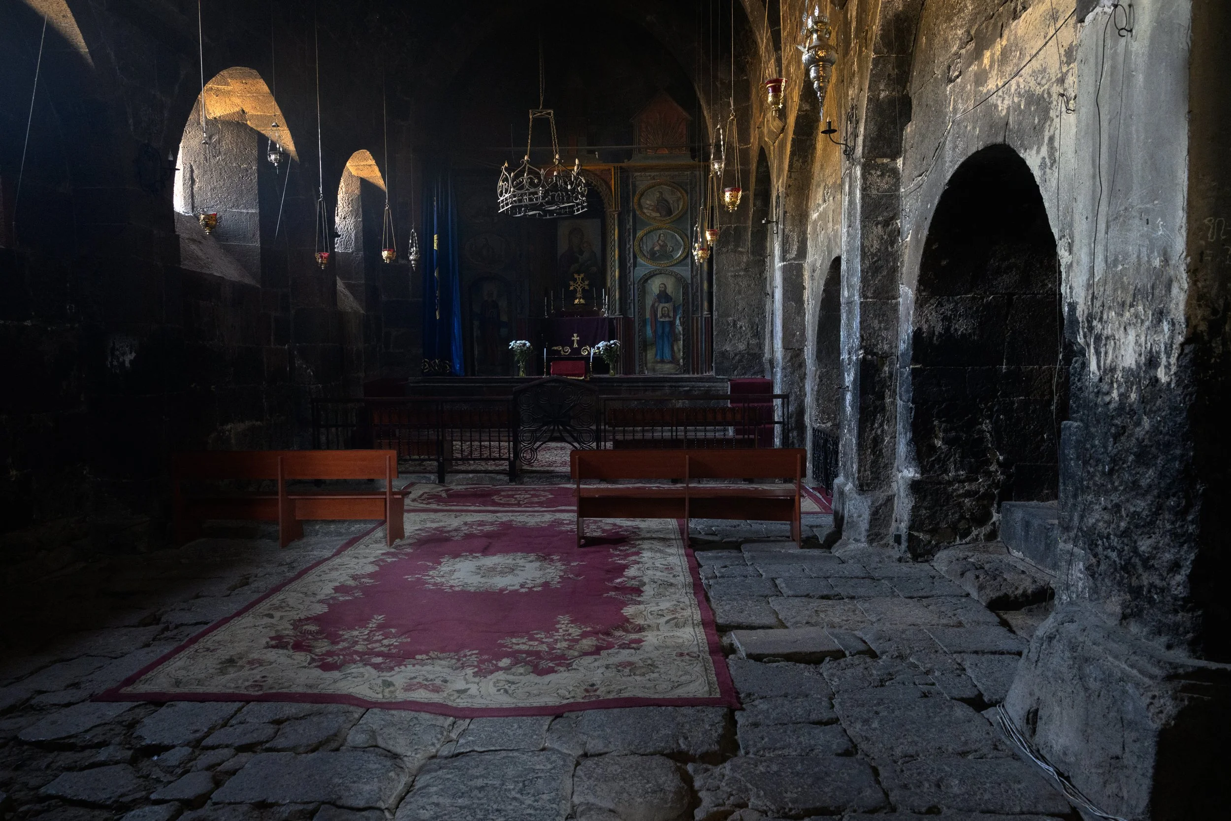 Interior of an old stone church with illuminated arched windows, a decorated altar, and wooden pews on a worn stone floor.