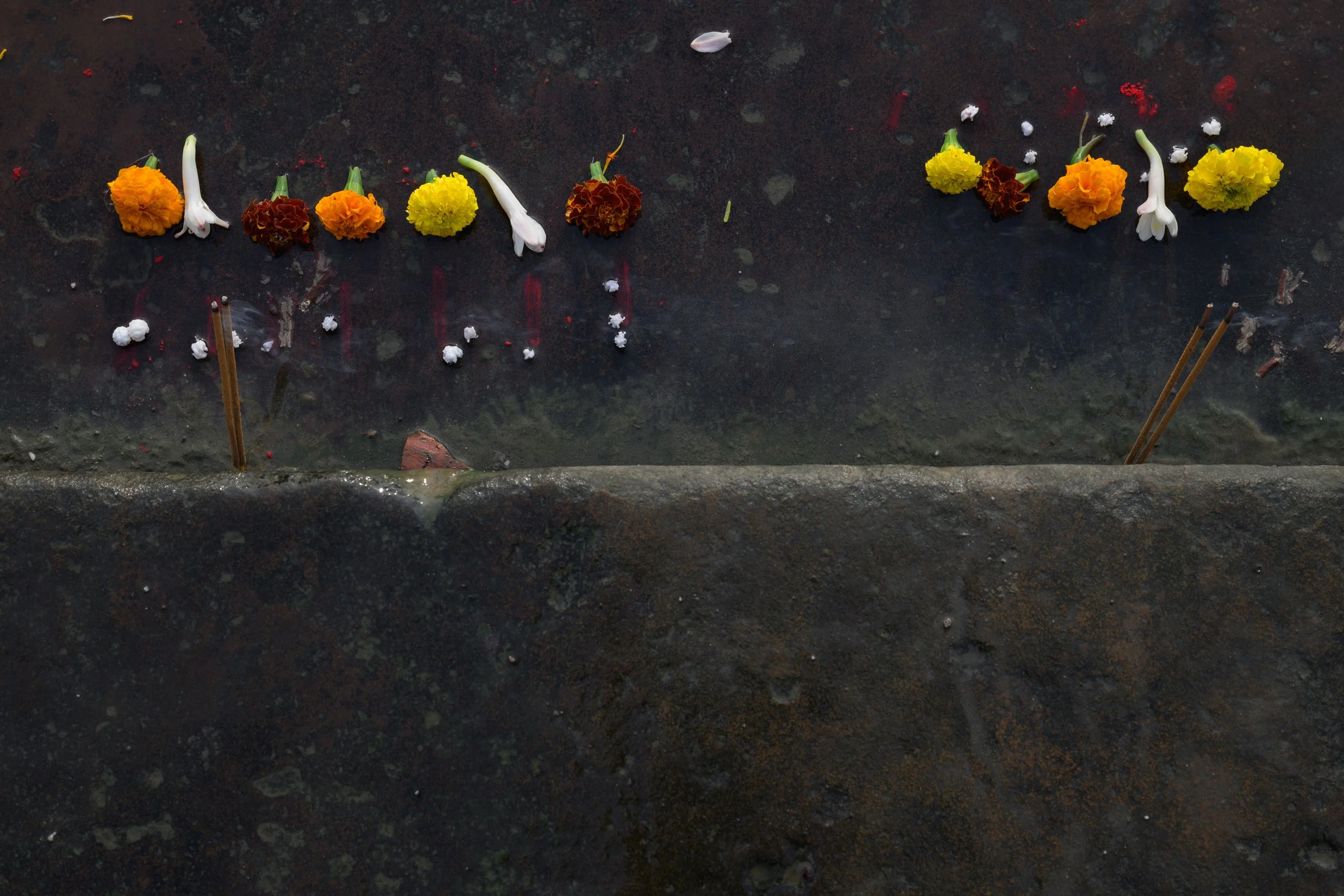 Flowers and white flowers with yellow and orange blooms are arranged on a stone surface, with incense sticks placed in the ground, suggesting a ceremonial or ritual setup.