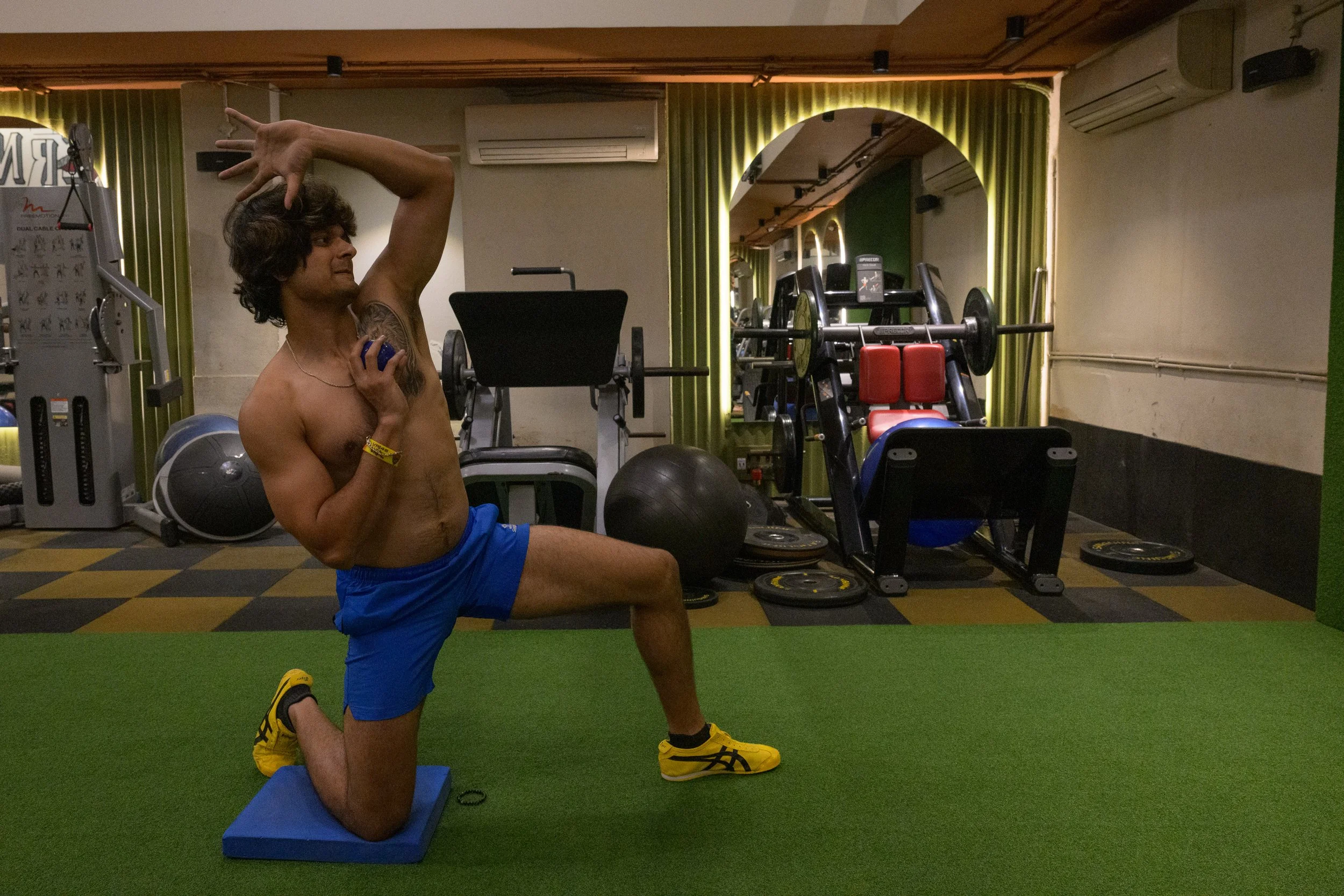A shirtless man in blue shorts and yellow athletic shoes performs a kneeling stretch on a blue yoga block on a green mat in a gym, holding a blue object in his right hand with his left hand raised above his head.
