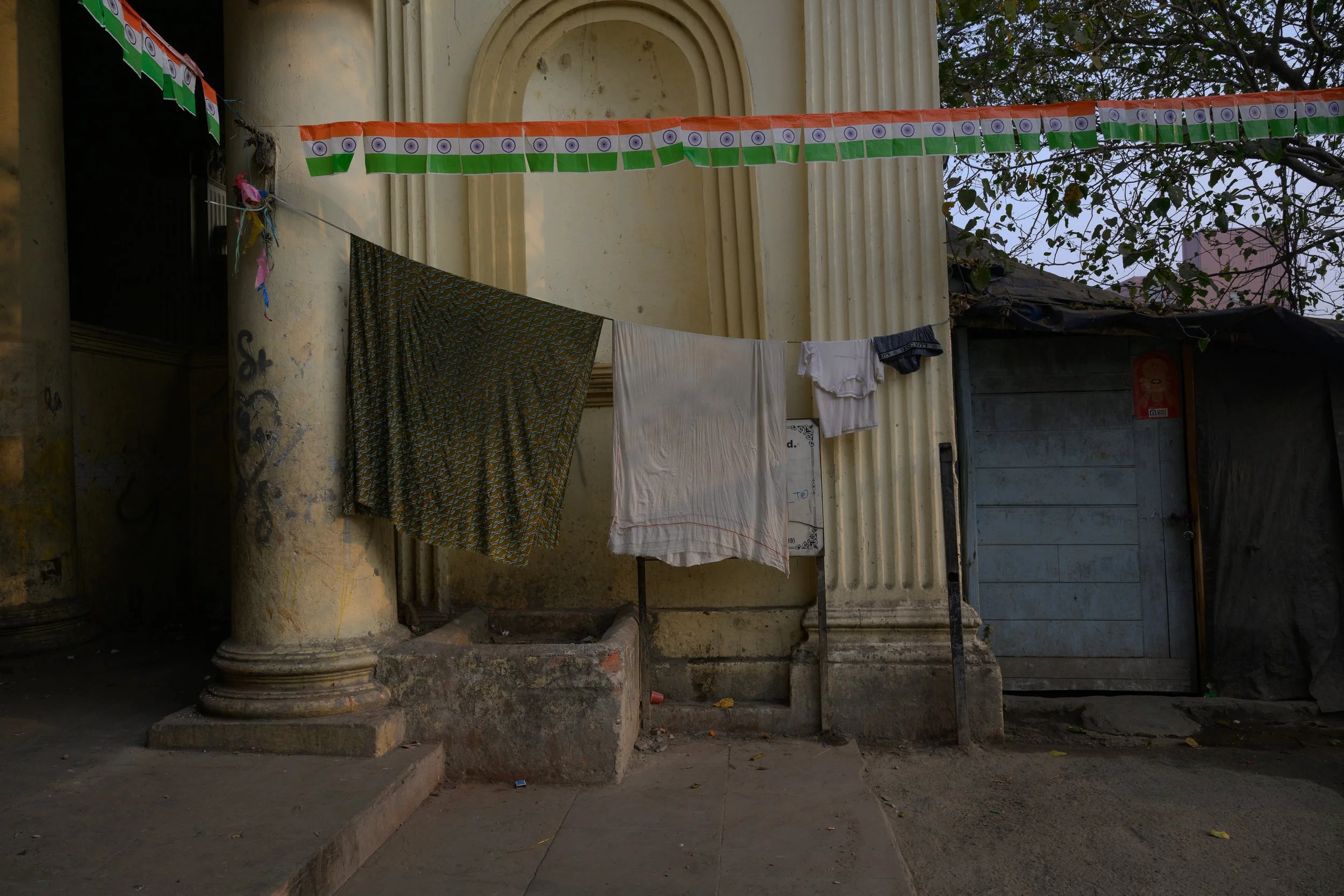 Clothes drying on a line in front of a weathered building with columns and an arch, with Indian flags strung above.