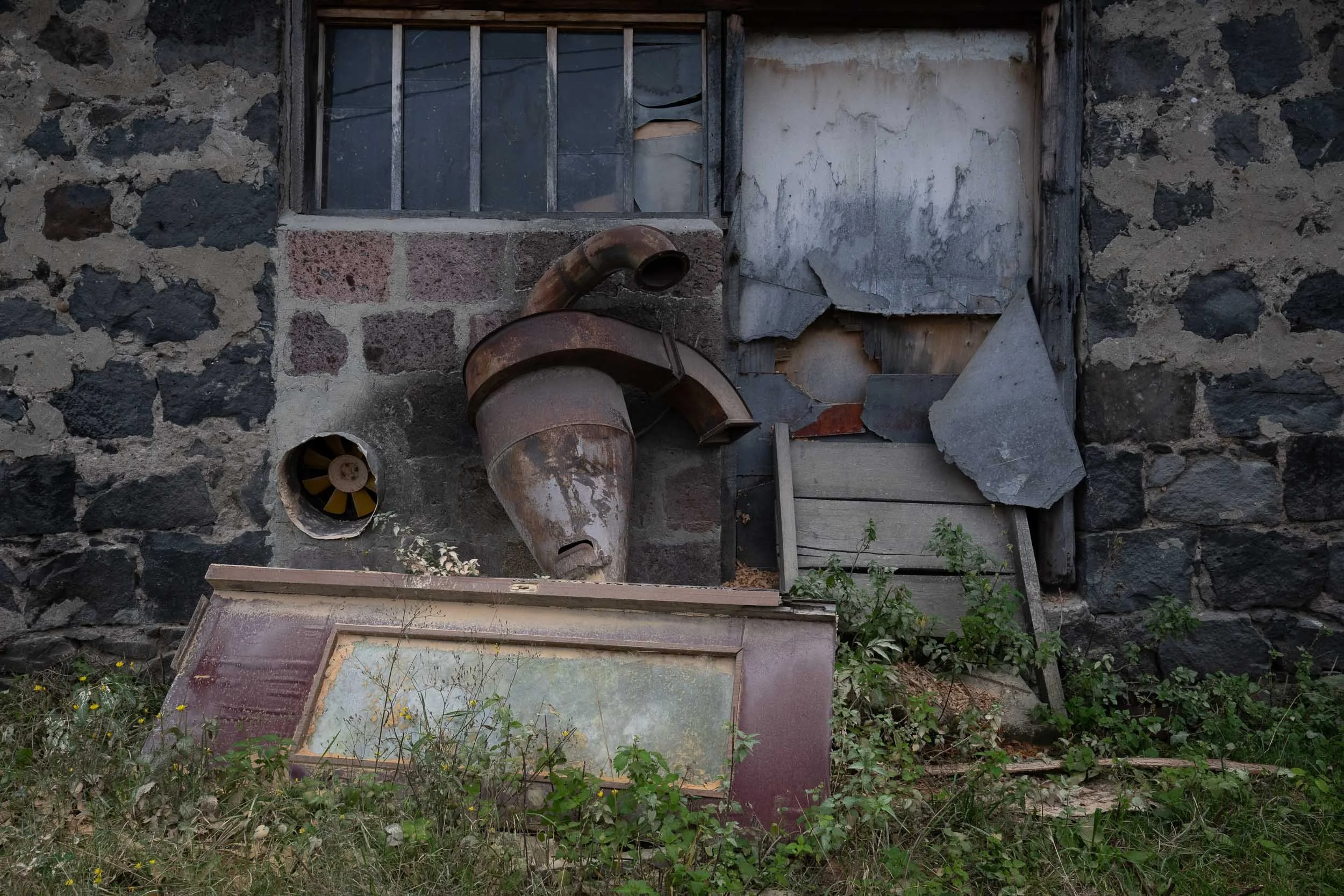 A weathered stone wall with a broken window and an old rusty metal chimney pipe. There is a glass window lying flat on the ground surrounded by overgrown grass and weeds.