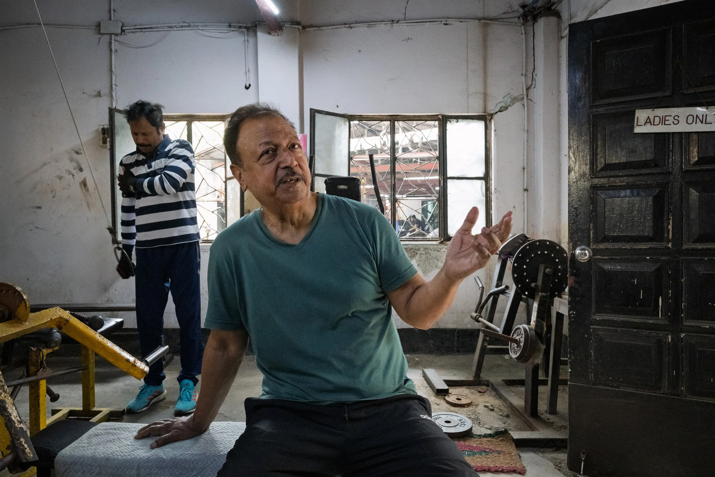 A man in a teal shirt sitting on a bench in a gym, gesturing with his right hand while speaking. Another man in a striped shirt is standing in the background near a window, looking down. The gym has various exercise equipment, and a 'LADIES ONLY' sig