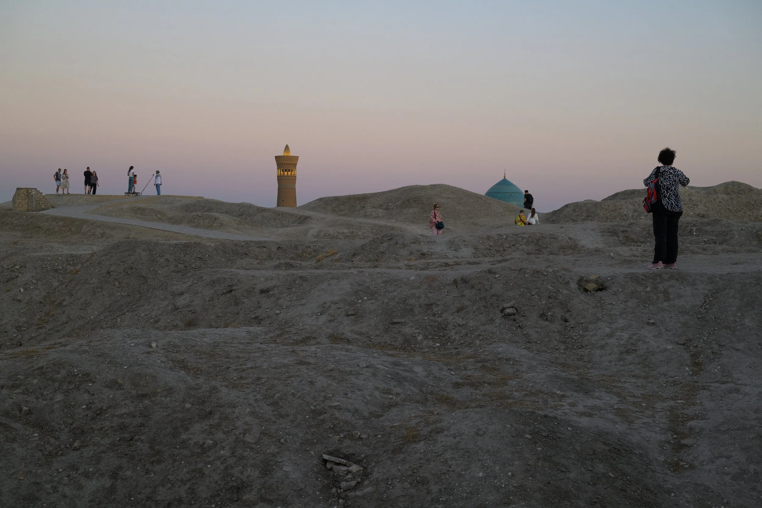 People walking and sitting on a barren, rocky landscape with historical Islamic architecture, including a minaret and a dome, under a pastel sky at dusk.