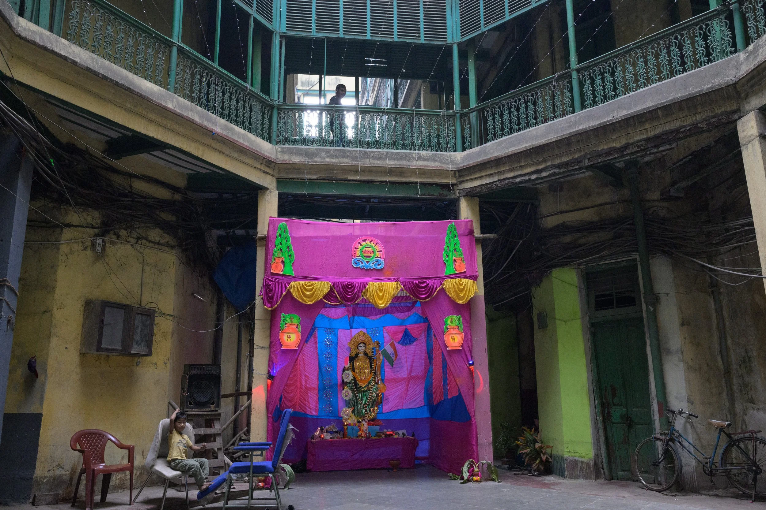 A temporary religious shrine decorated with pink and purple fabric, featuring a statue of a deity adorned with flowers, in an open-air courtyard surrounded by yellow and green walls. There are chairs and bicycles nearby, and a child is sitting on one