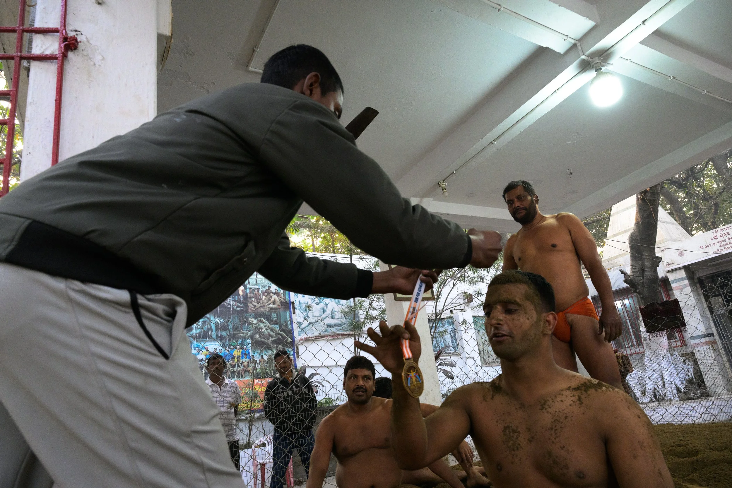 A man in a gray jacket and gray pants is handing a medal to a shirtless wrestling contestant covered in dirt, seated on the ground. Another shirtless man stands behind him, also dirty, with a few people and a fence in the background.