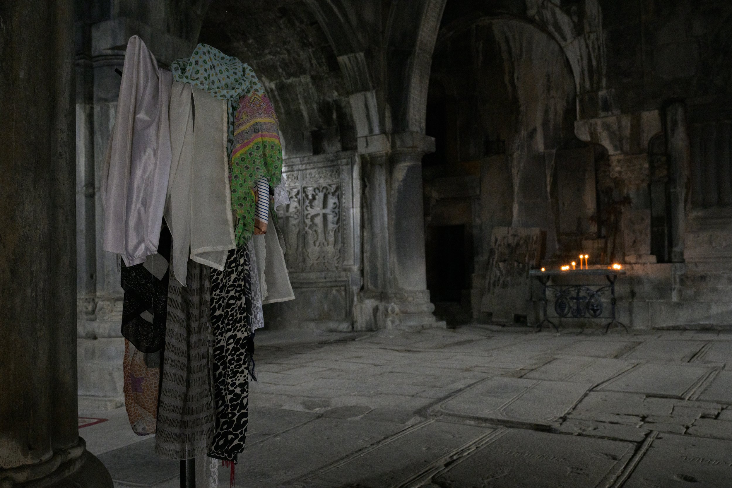 Clothing display with assorted colorful garments on a stand inside a dimly lit, ancient stone building with intricate carvings and a table with lit candles in the background.