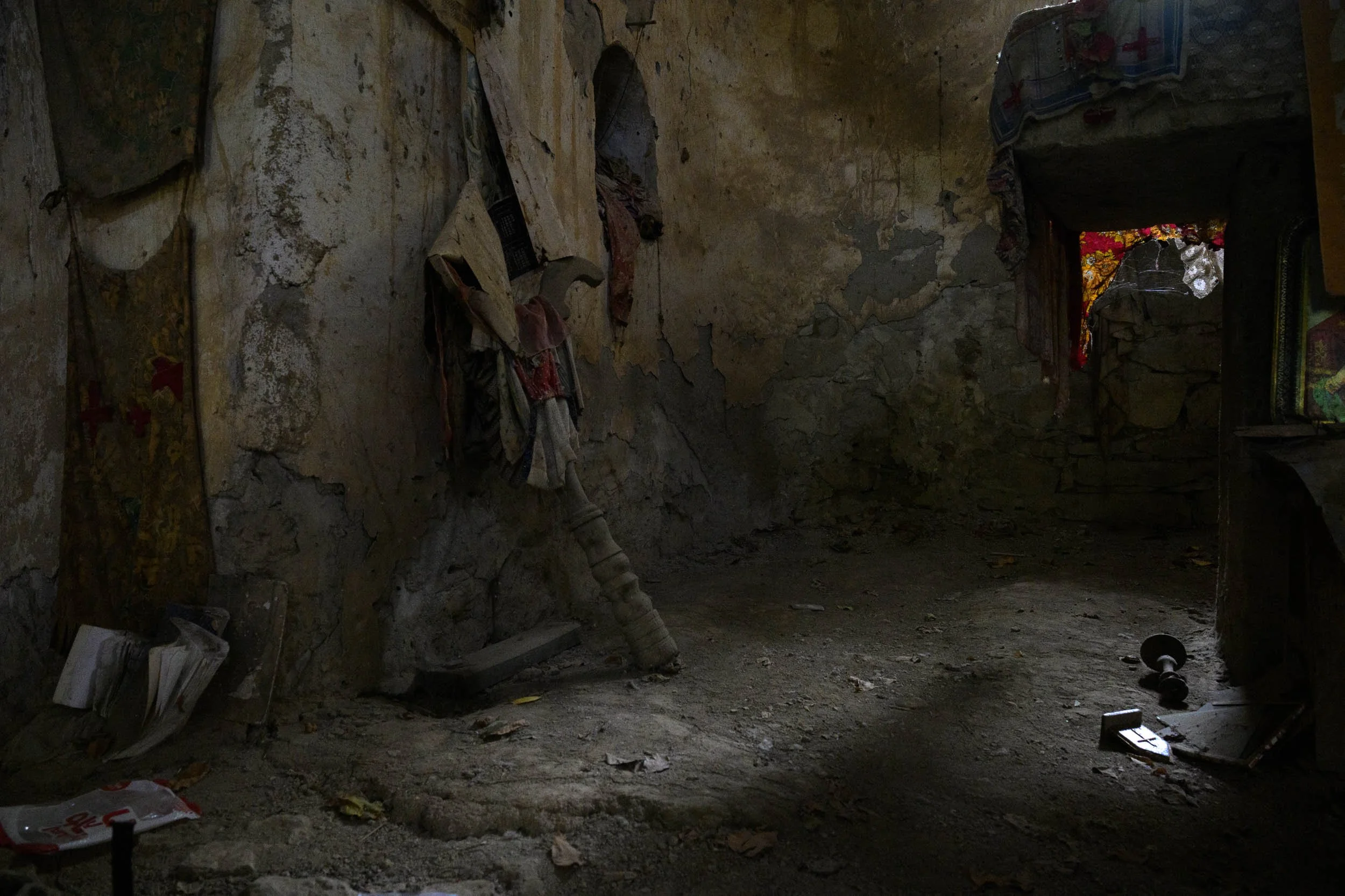 An empty, dark room with bare and damaged stone walls, scattered debris, and a frayed flag hanging from the wall. There are old shoes on the floor and some strange objects, with a small illuminated area showing a glimpse of red, yellow, and floral pa