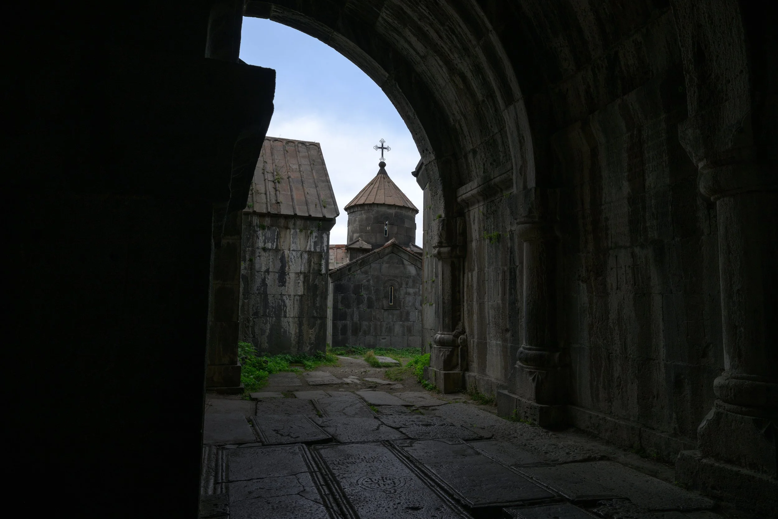 View through a dark stone archway of an old church with a small dome topped with a cross, stone walls, and green grass outside.