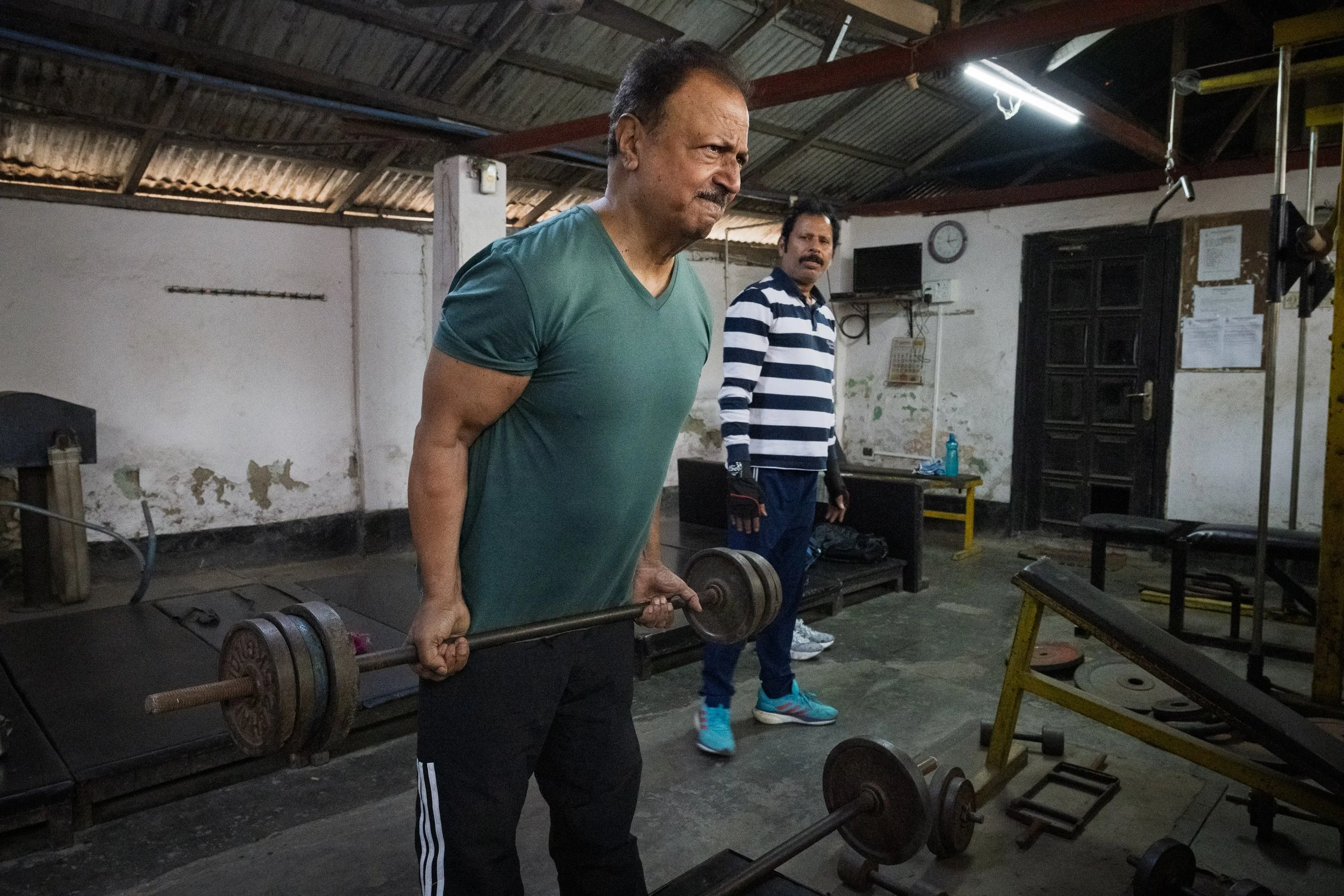A man lifting a barbell in a gym with another man standing behind him.