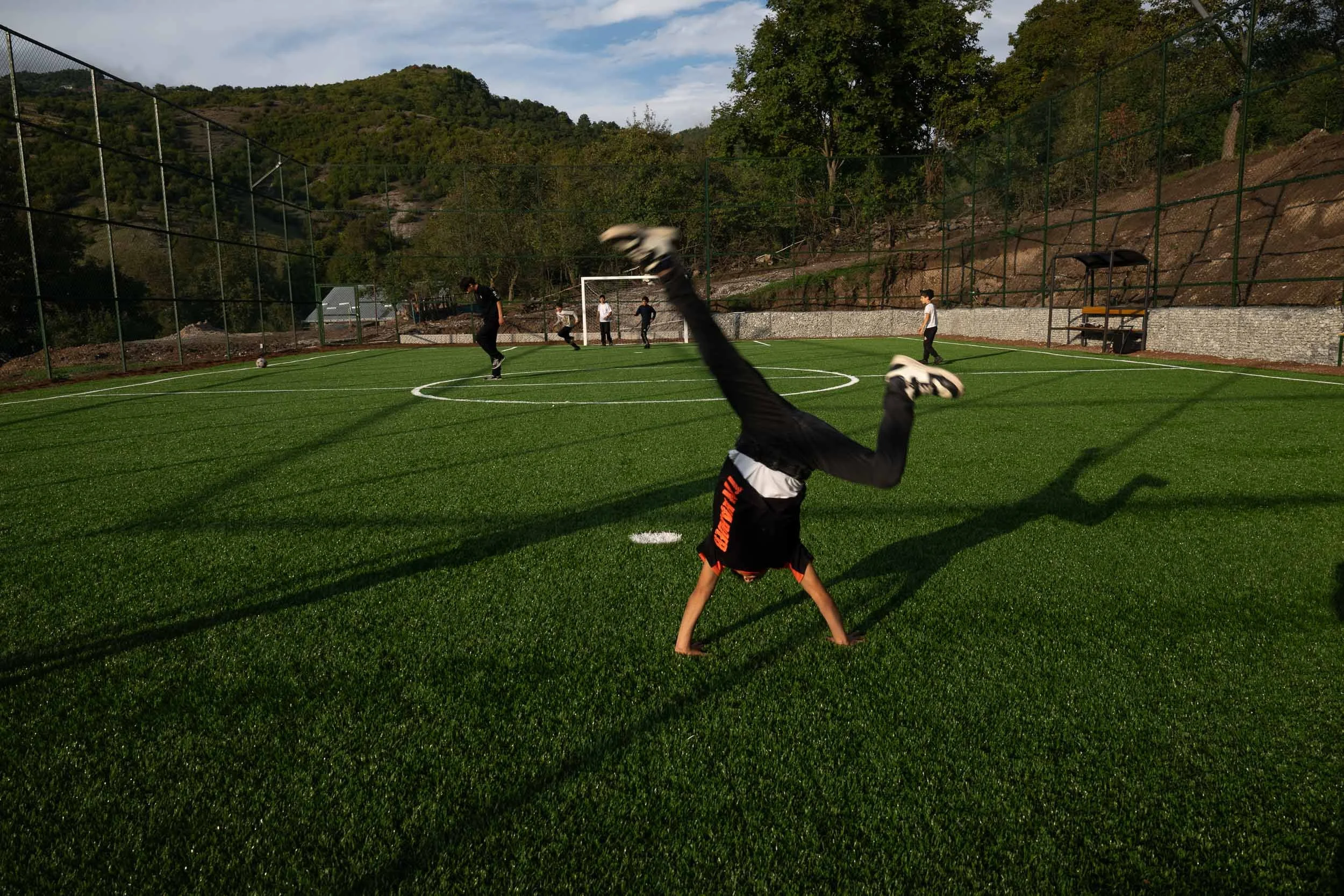 Young boy performing a handstand on a football field with other kids playing soccer in the background, surrounded by hills and trees.